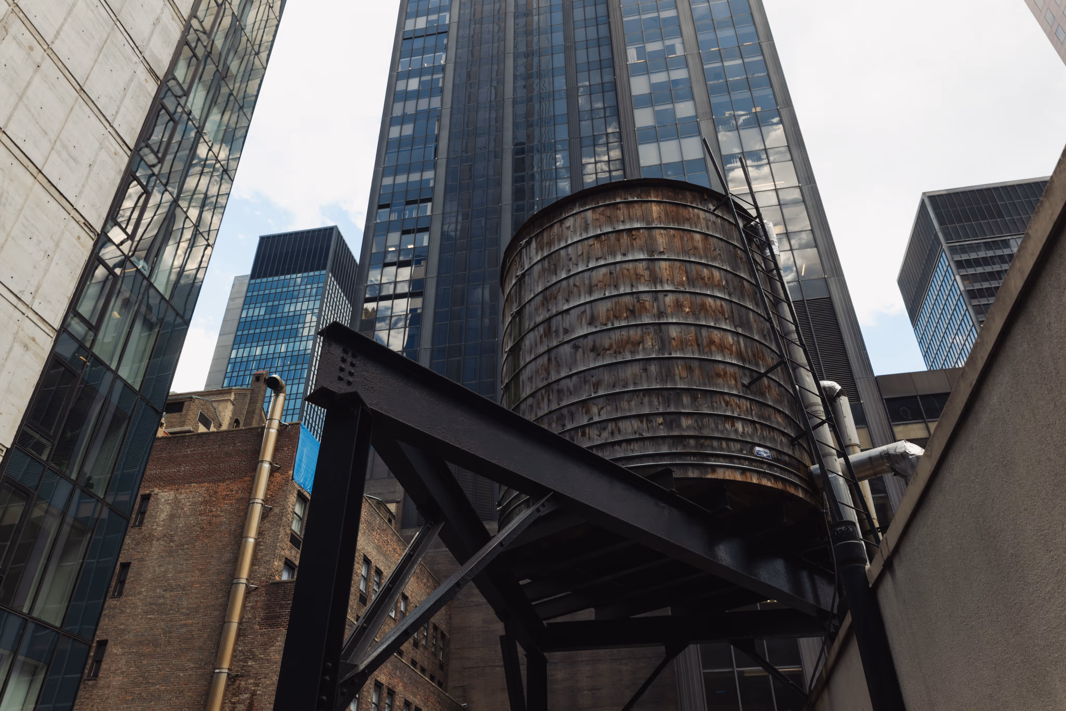 Wooden rooftop water tank supported by black metal framework against backdrop of tall city skyscrapers.