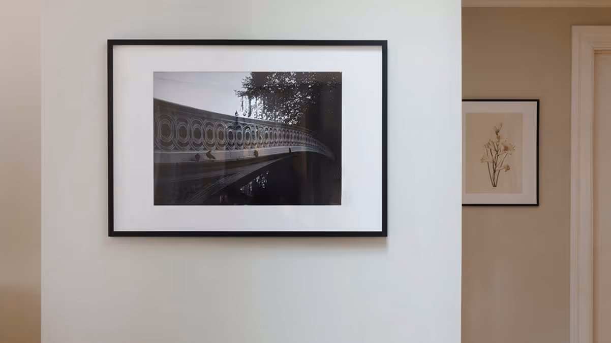 Wicker bench under a black-and-white framed photo of a bridge on a white wall with hardwood floor.
