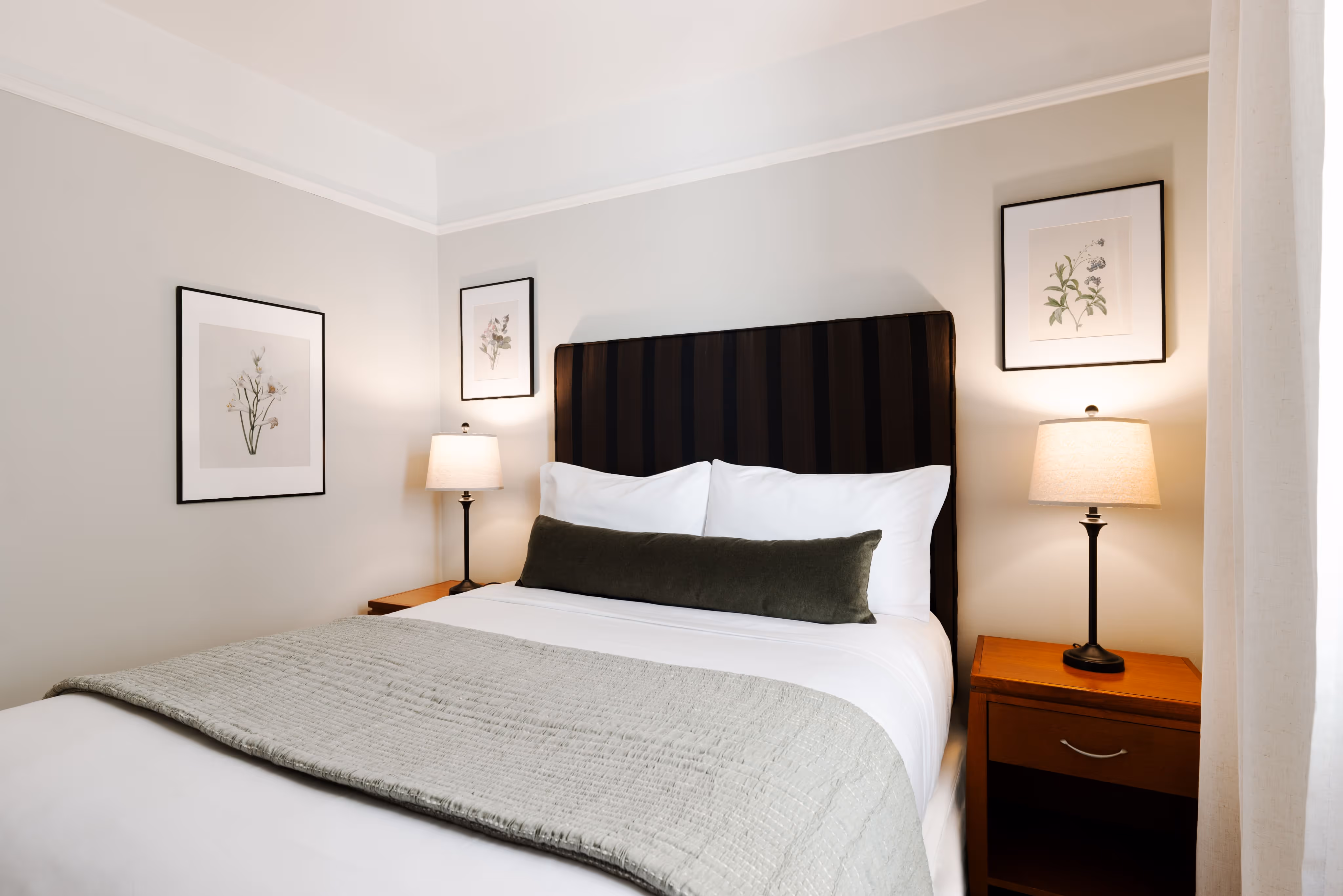 Minimalist bedroom with a queen bed featuring white linens, a long green bolster pillow, striped dark headboard, two bedside tables with lamps, and framed botanical prints on beige walls.