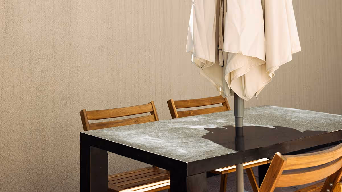 Outdoor patio with a black table, wooden chairs, and a closed beige umbrella against a cityscape background.