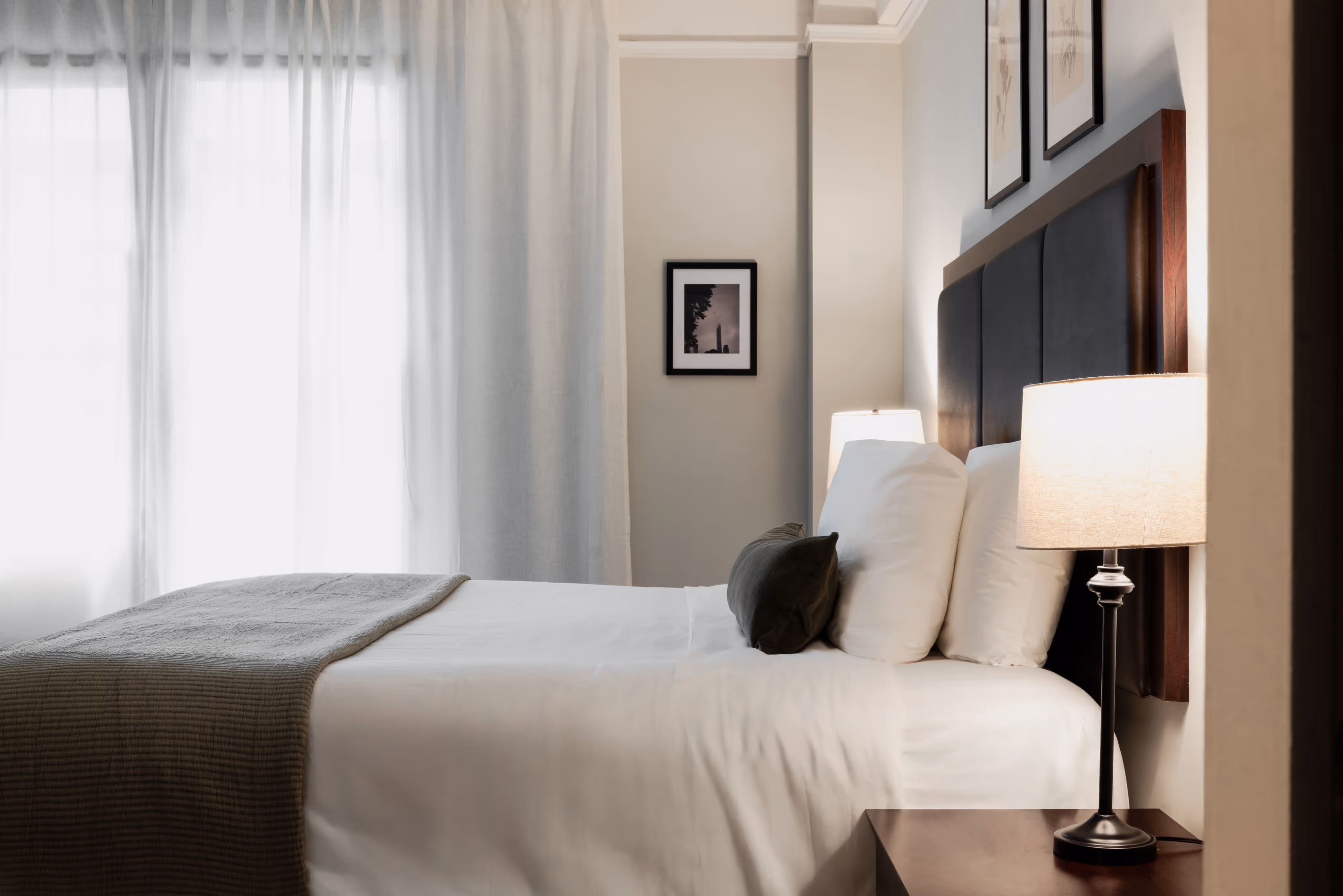 Minimalist bedroom with a neatly made bed, white linens, gray blanket, and two lamps on either side.