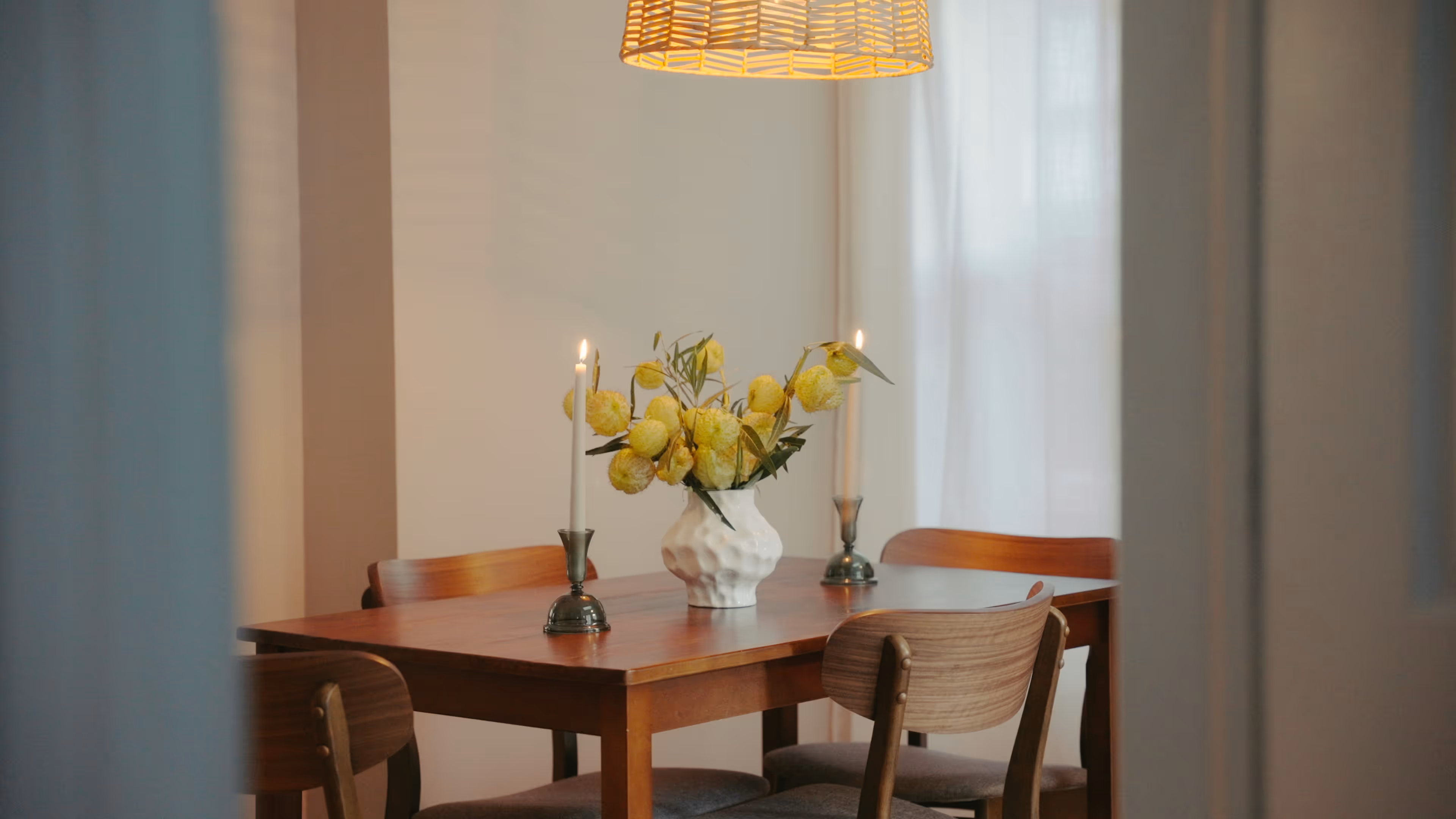 Wooden dining table with a white vase of yellow flowers and two lit candles in brass holders under a wicker pendant light.