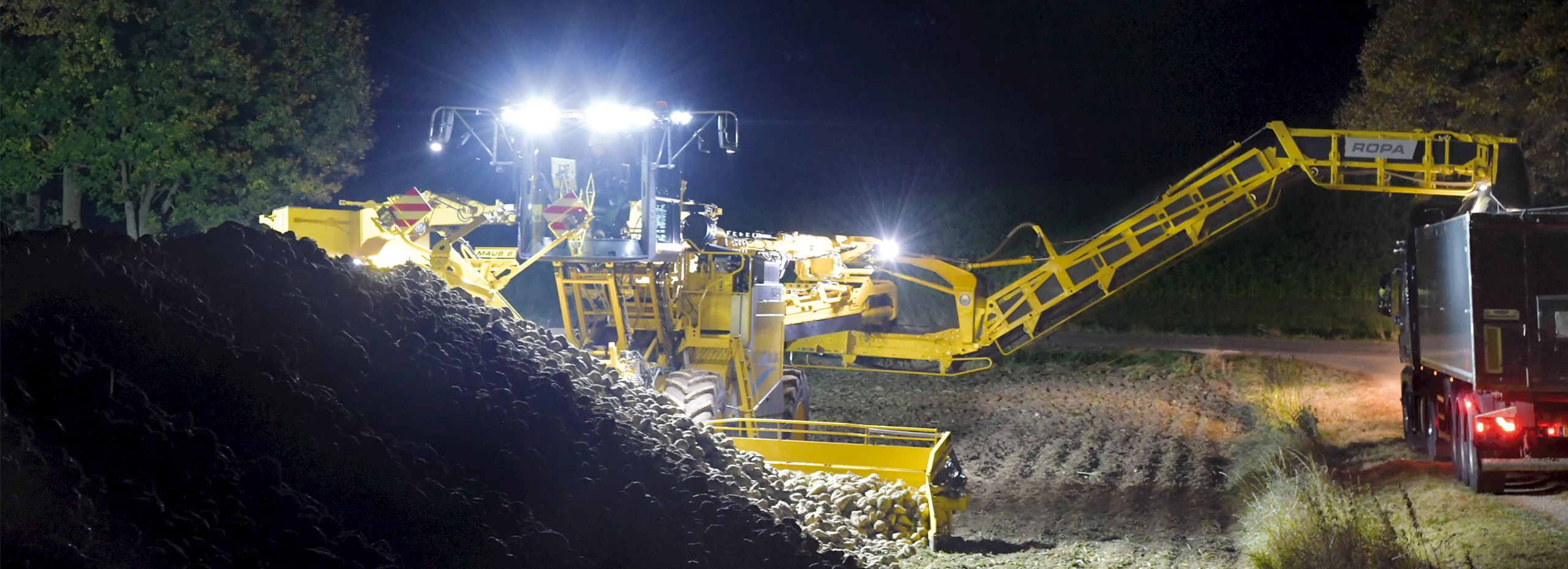 A ROPA Maus 6 with bright, LED lights loading sugar beets from a field pile onto a truck at night