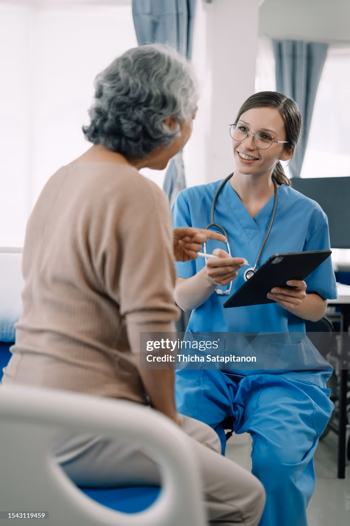 Eine Ärztin spricht mit älterer Patientin auf Augenhöhe und hält ein Tablet; Beratung im Klinik- oder Pflegeumfeld.