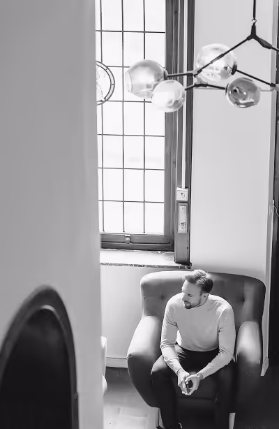 Jochen sitting on a cushioned armchair near a window, under a modern chandelier with glass spheres in the Ghent office.
