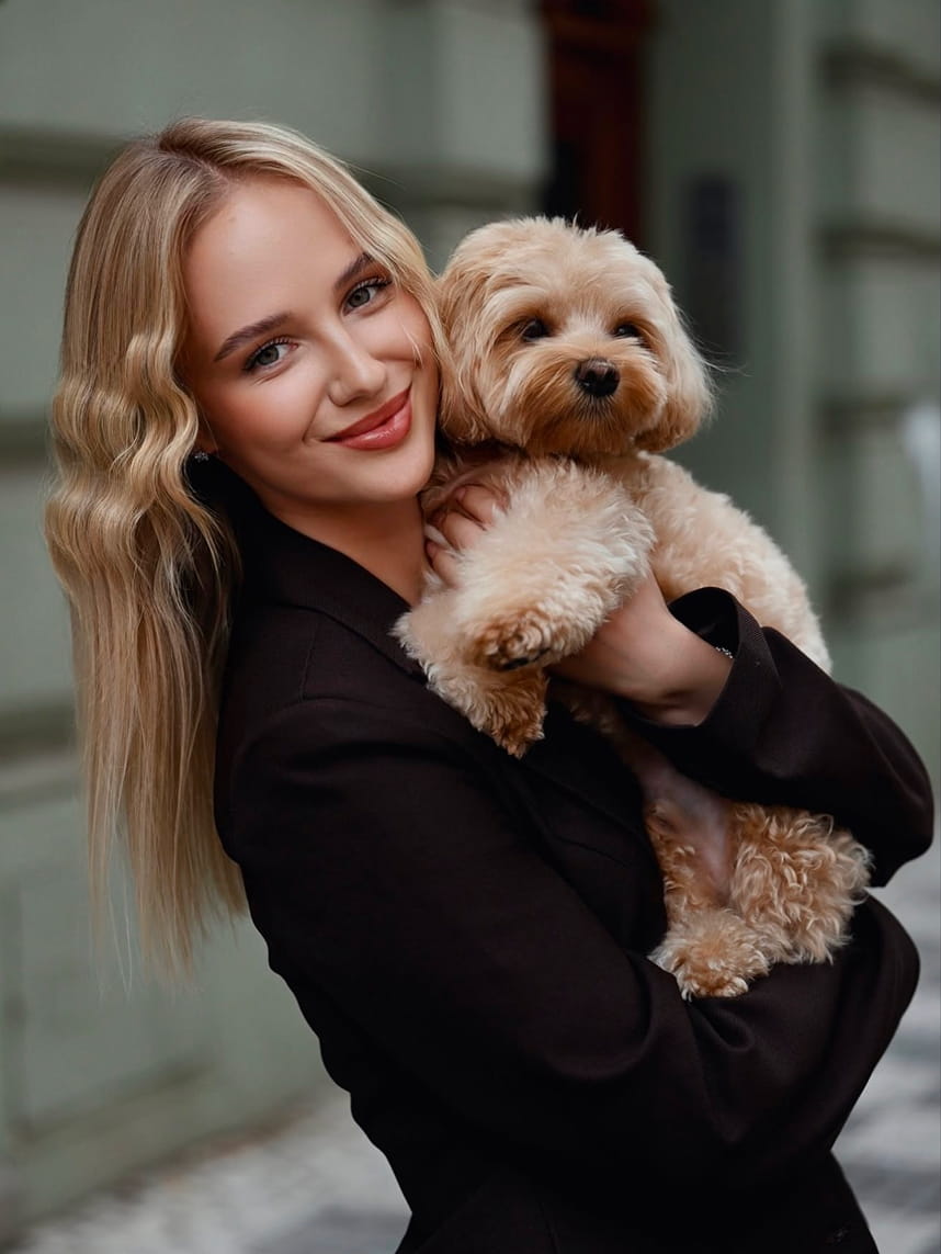 Smiling blonde woman wearing a black jacket holding a small fluffy light brown dog.