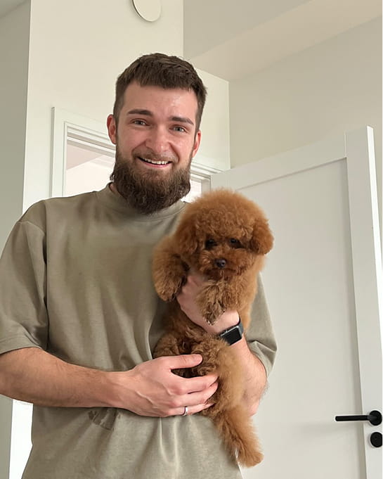 Smiling man with a beard holding a small fluffy brown dog indoors.