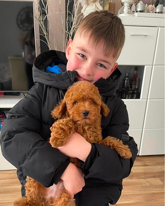 Young boy wearing a black jacket hugging a curly brown puppy indoors.