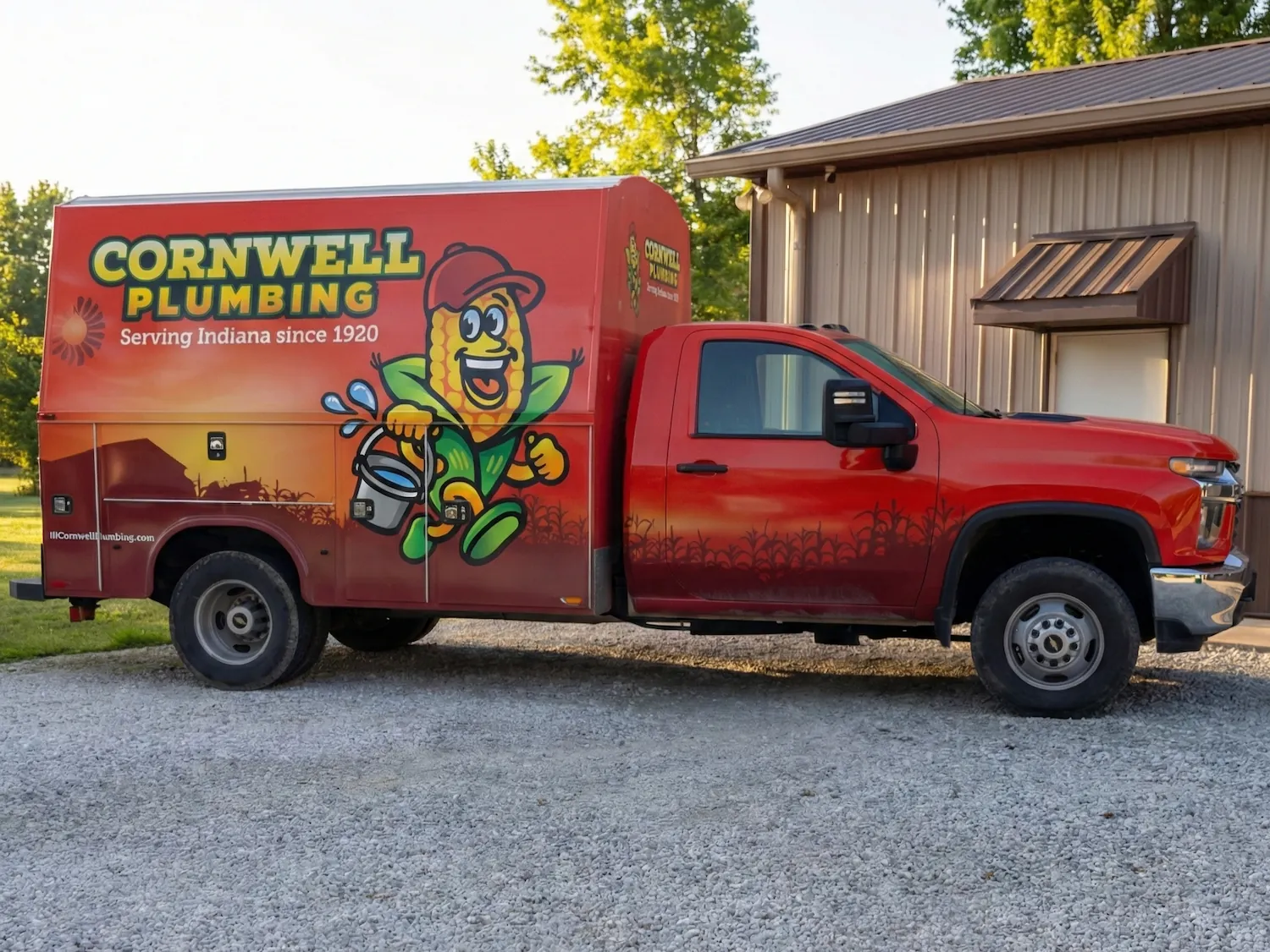Red Cornwell Plumbing service truck with a cartoon corn mascot holding a bucket, parked on gravel near a building.