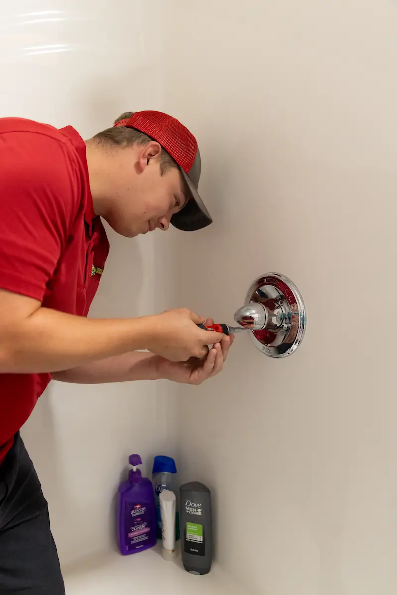Technician in a red shirt and cap repairing a shower handle with a screwdriver in a bathroom corner with bath products.