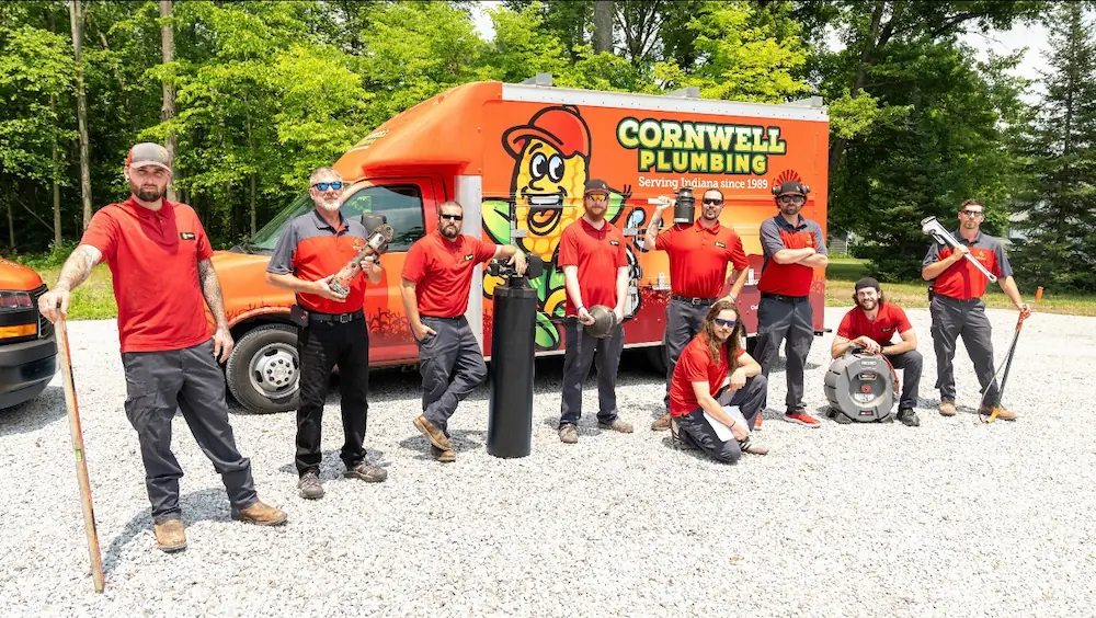 Nine Cornwell Plumbing workers in red shirts posing with plumbing tools and equipment in front of an orange company truck.