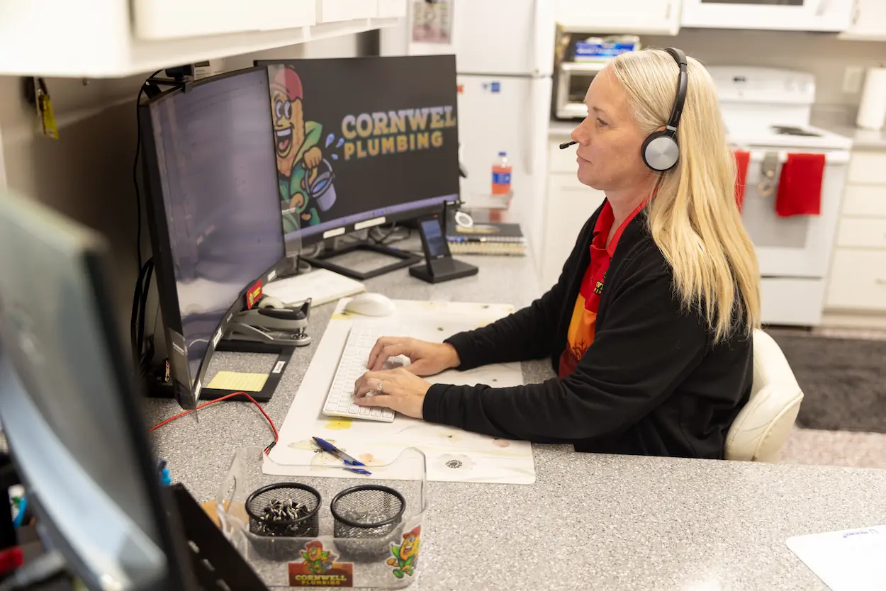 Woman wearing a headset typing on a keyboard at a desk with dual monitors displaying Cornwell Plumbing logo.