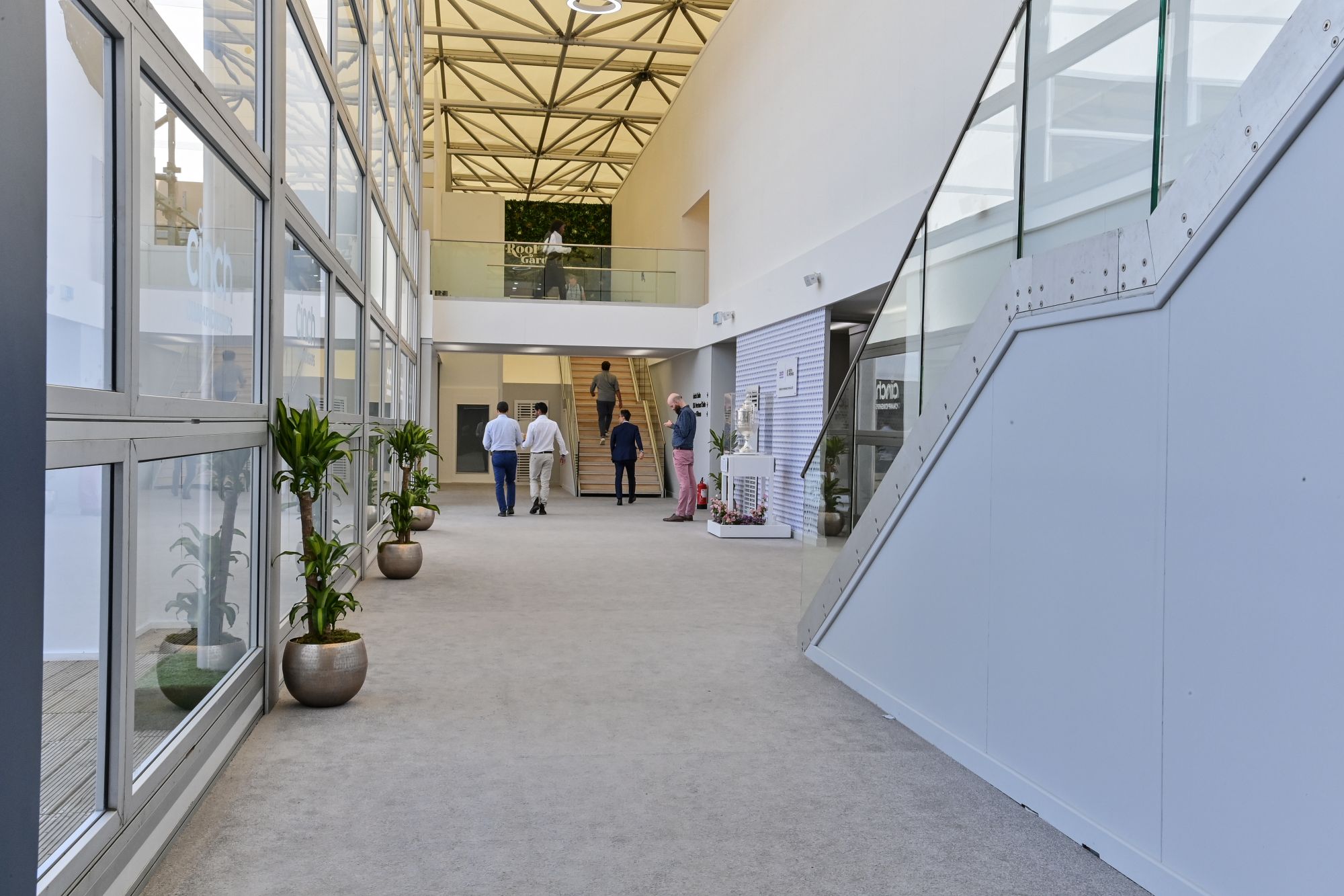 Modern indoor walkway with large windows, potted plants, stairs, and people walking and standing.