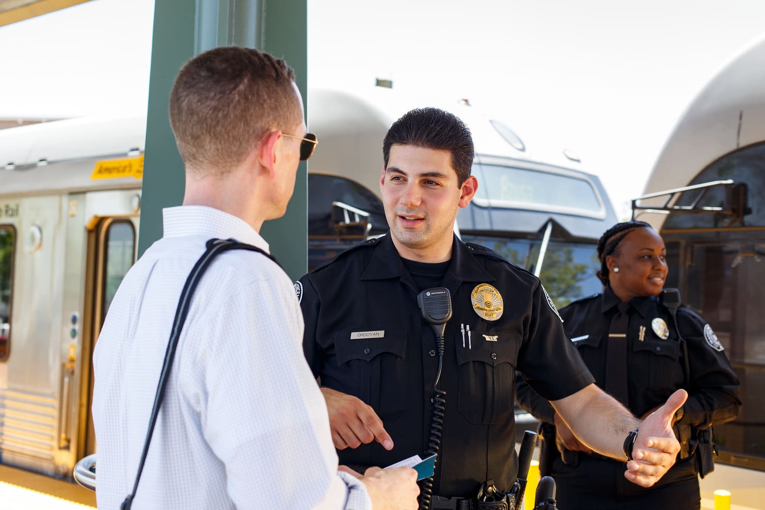 Two police officers talking to a man holding a ticket at a train station platform with a train in the background.