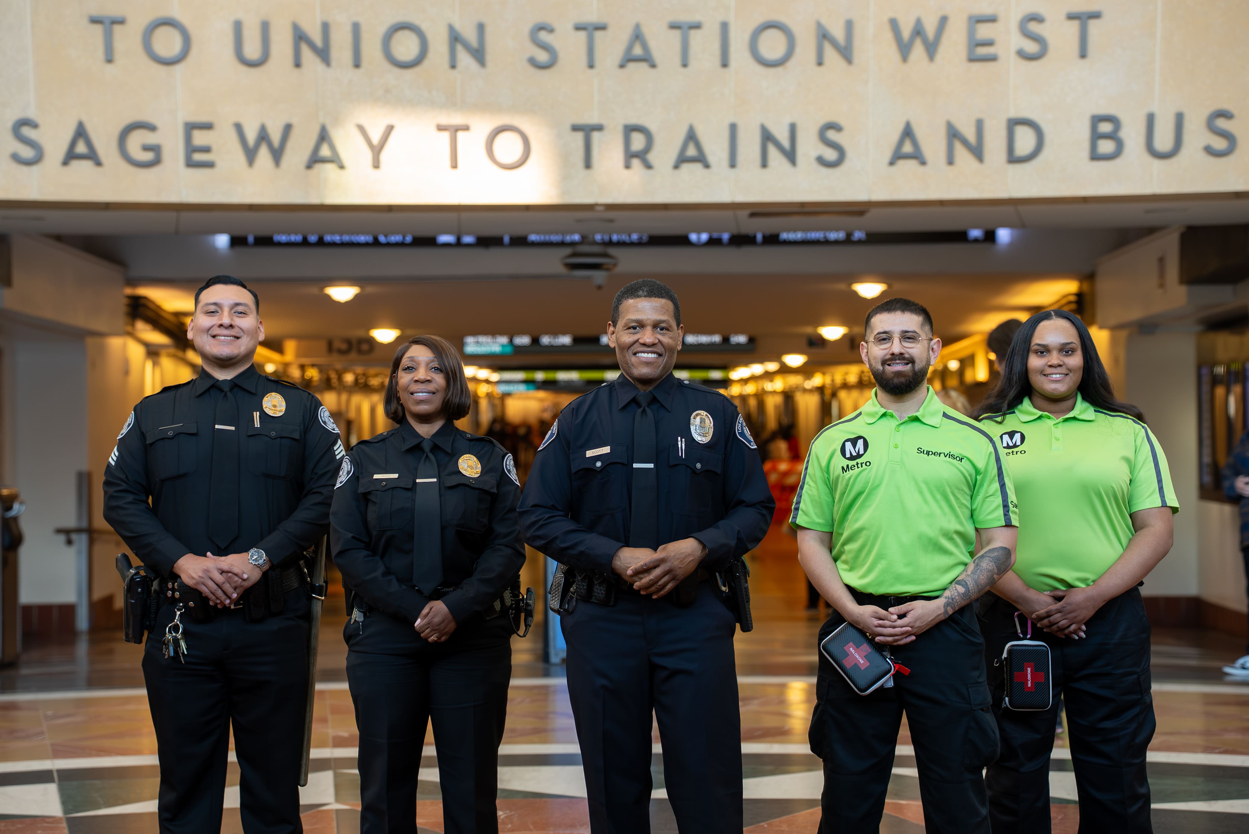 Three police officers in black uniforms and two Metro supervisors in lime green shirts standing inside a transit station under a sign reading 'To Union Station West Sageway to Trains and Bus'.
