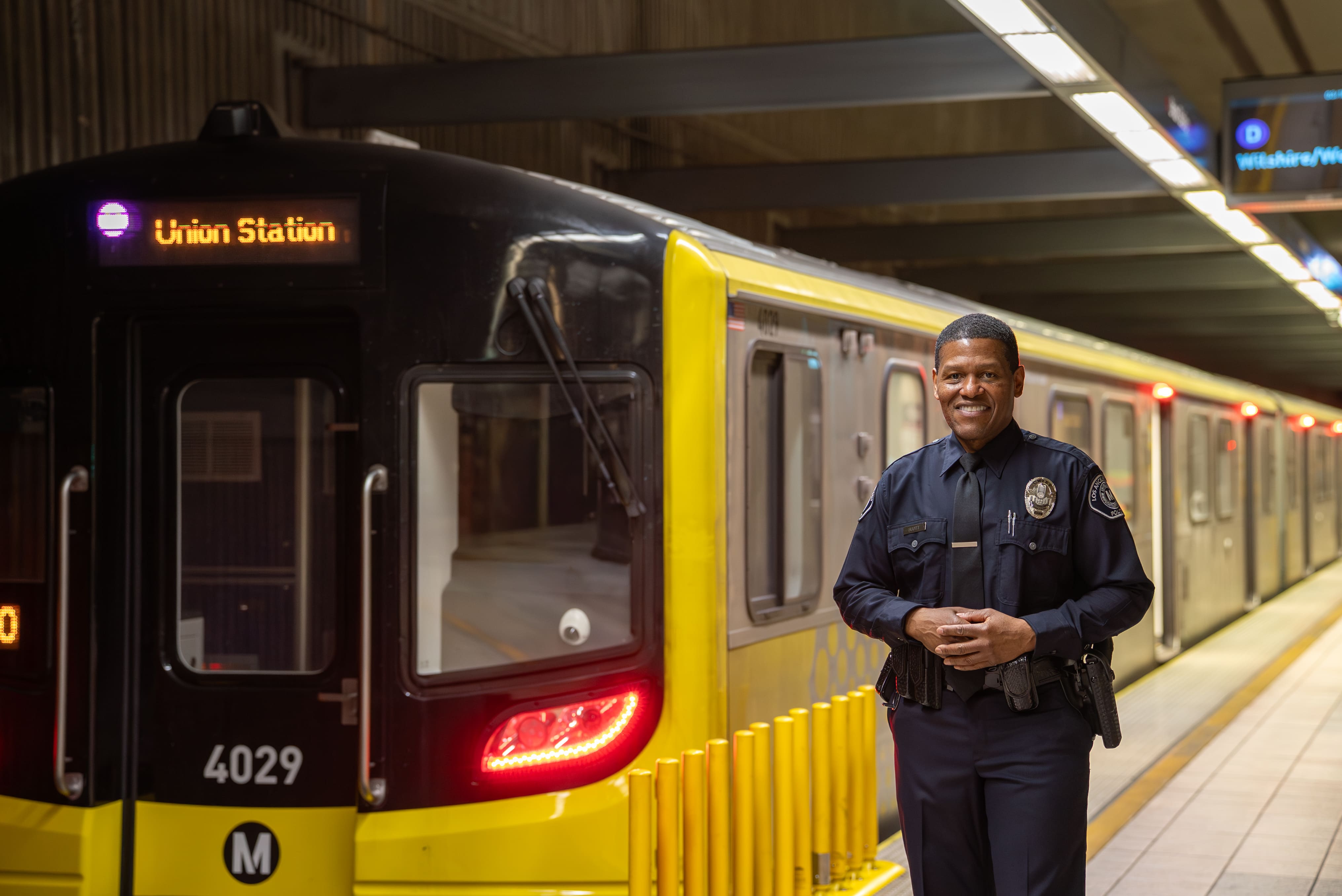 Smiling Los Angeles Metro police officer standing in front of a yellow subway train at a station with a Union Station sign.