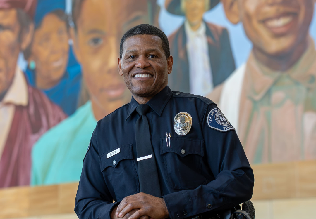 Smiling African American male police officer in uniform with badge and name tag 'Scott' sitting in front of a colorful mural.