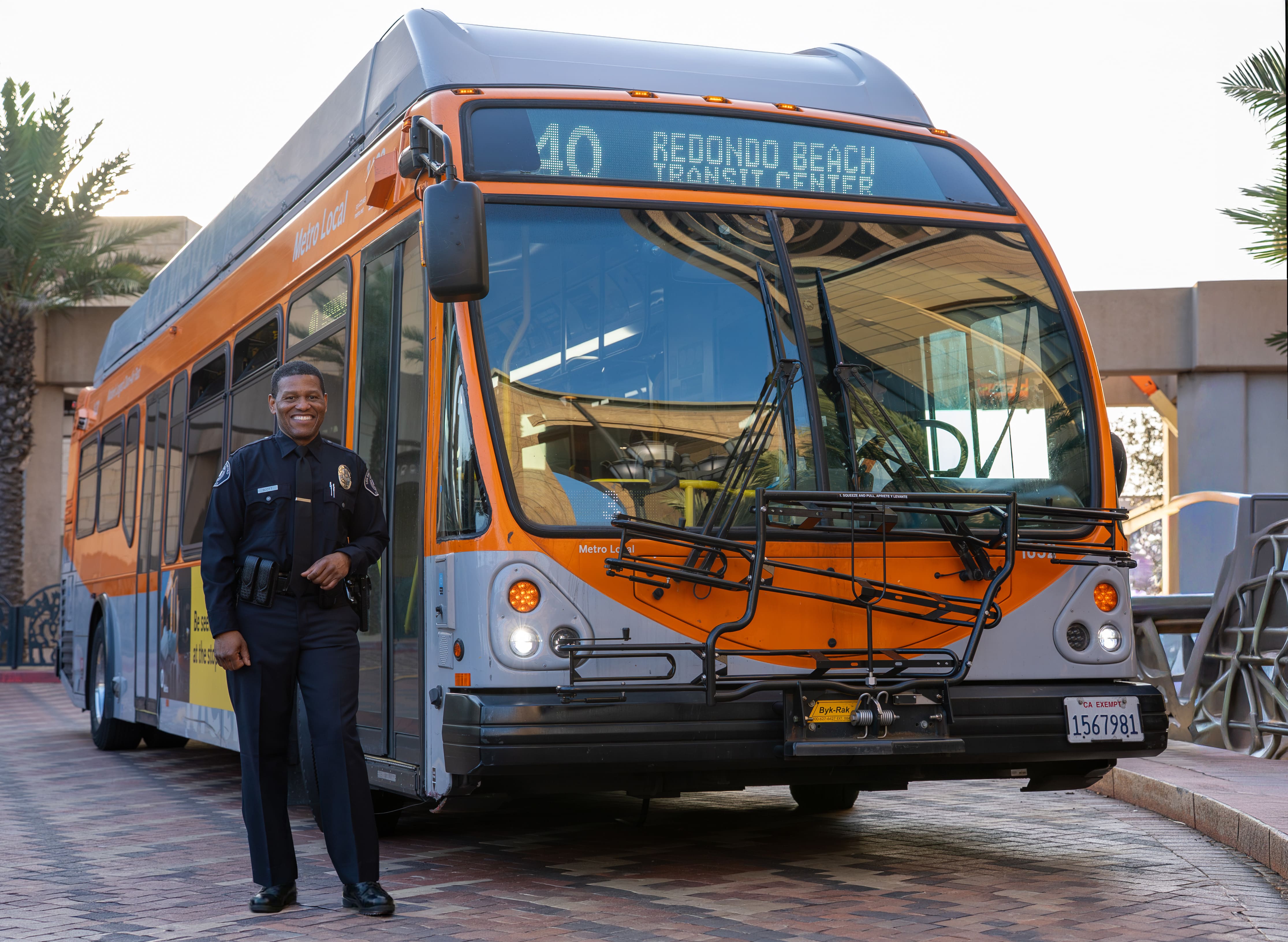 Uniformed transit police officer standing and smiling beside an orange Metro Local bus with destination Redondo Beach Transit Center.