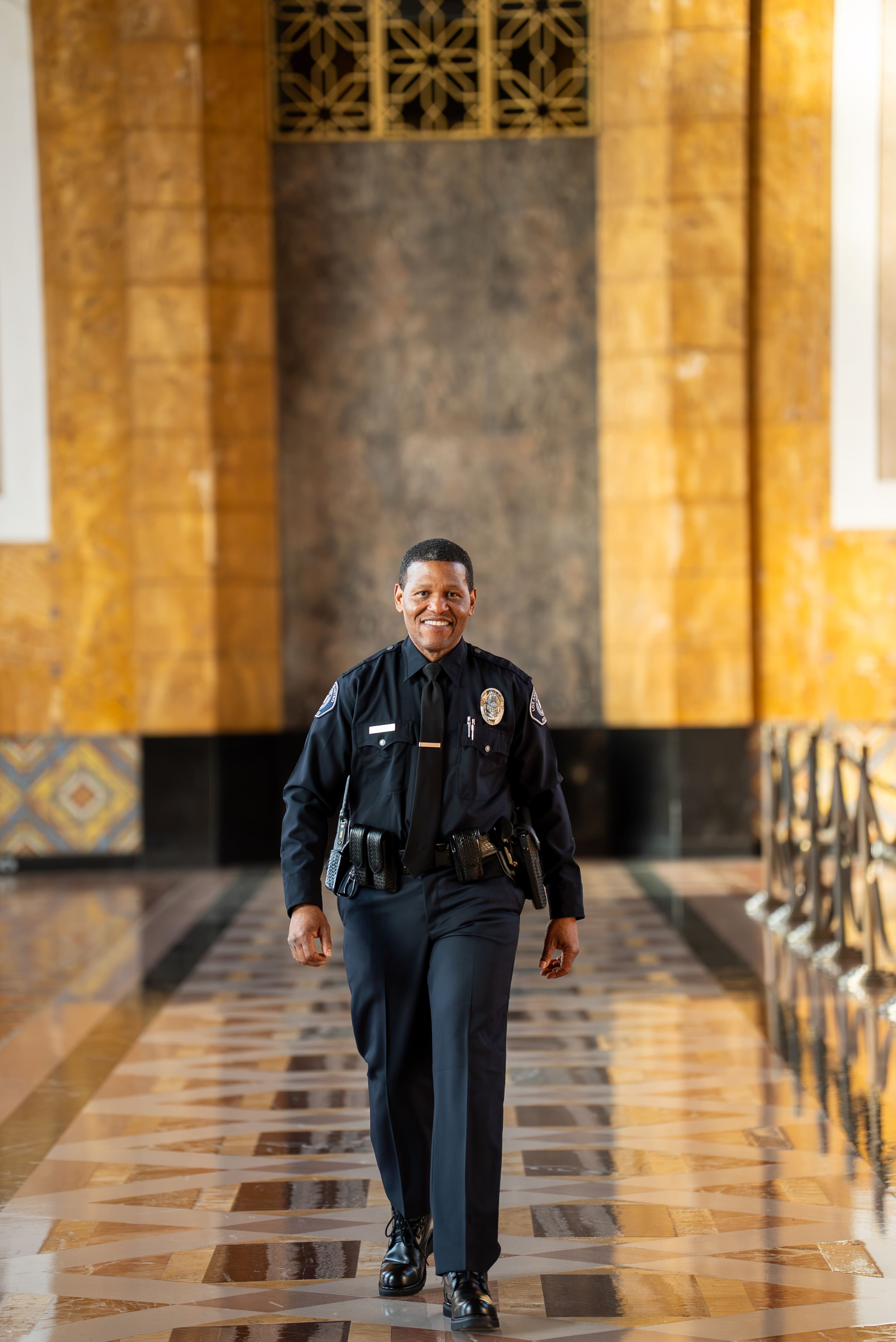 Smiling uniformed police officer walking through a hallway with patterned floor and golden walls.