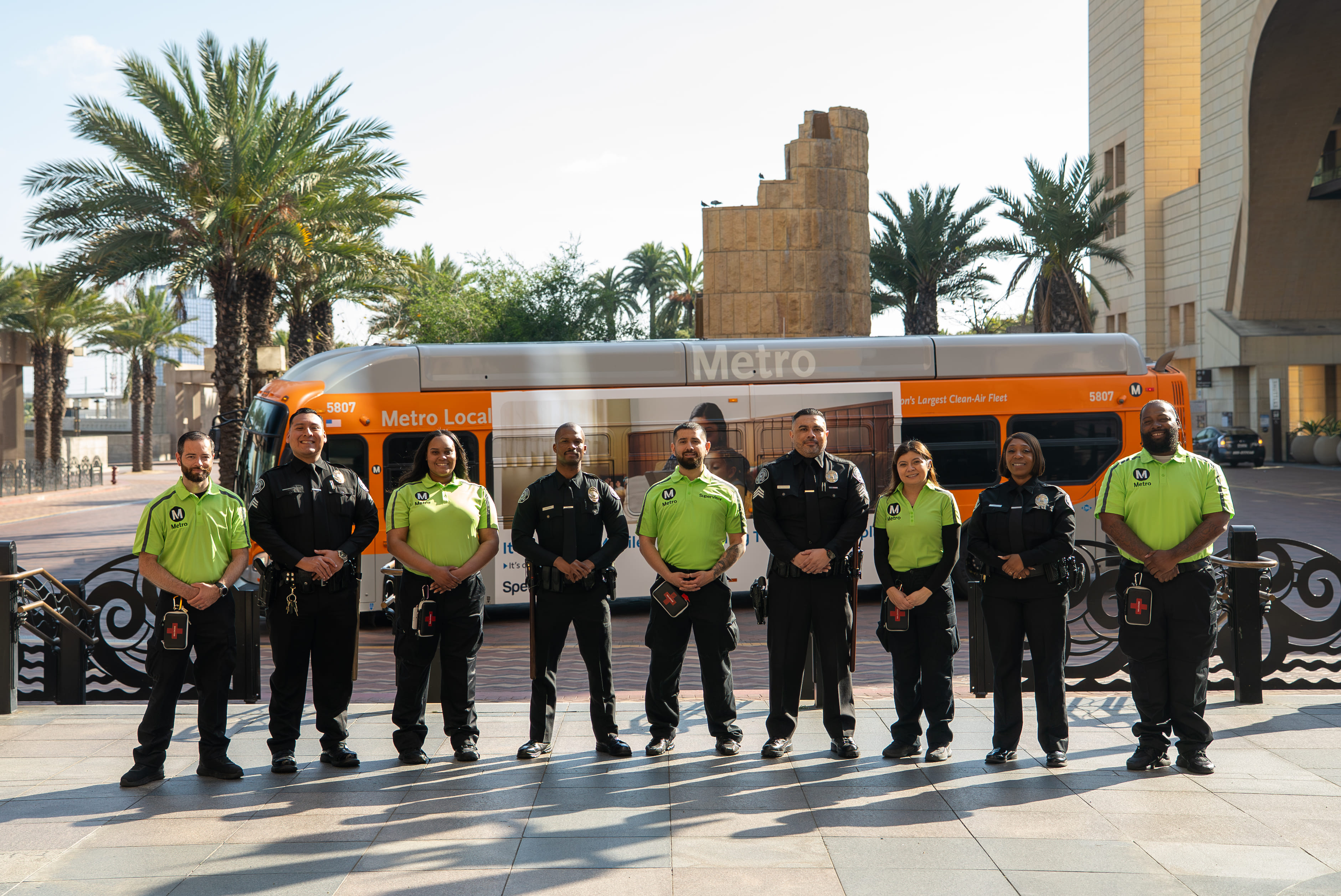 Group of nine LA Metro security officers and personnel standing in front of an orange Metro Local bus with palm trees in the background.