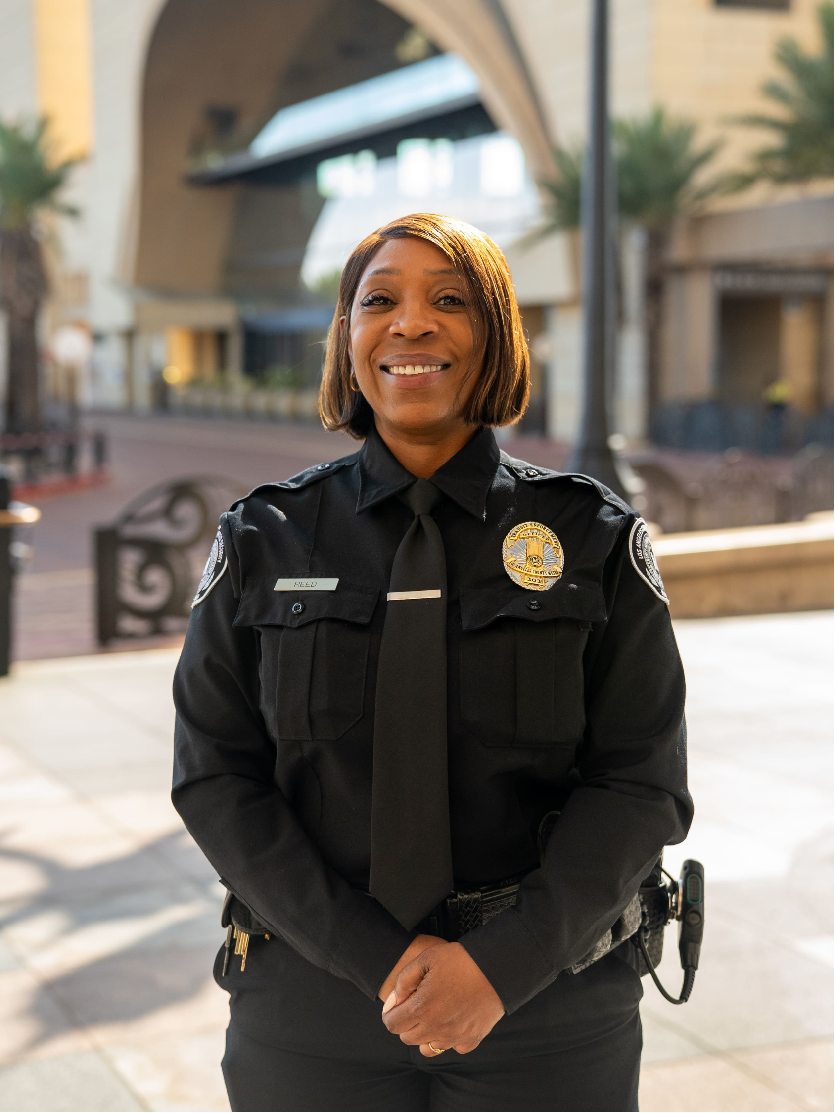 Smiling female security officer in black uniform with badge, name tag 'Reed', and radio equipment.