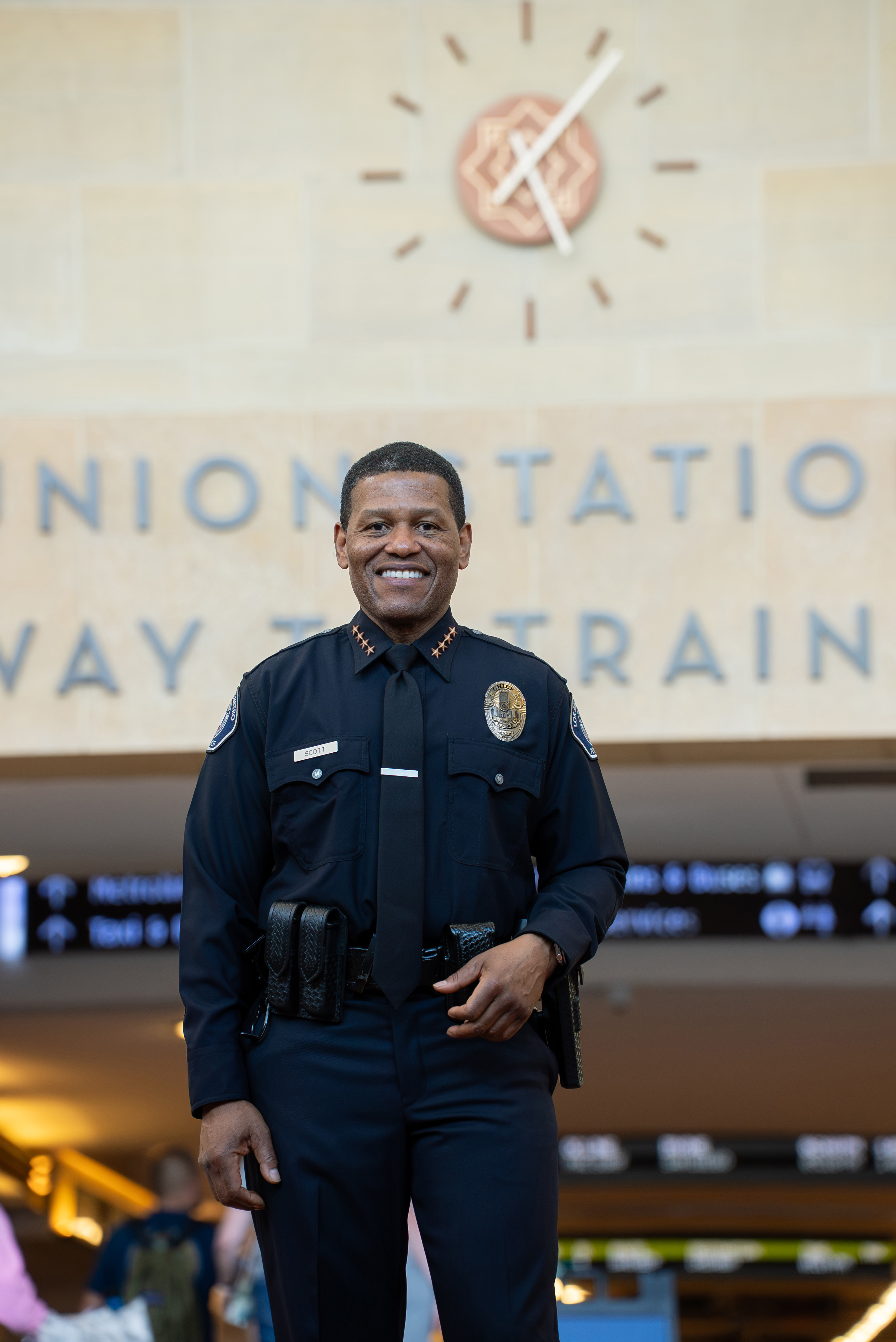 Uniformed police officer named Scott smiling inside a train station with a clock and signage in the background.