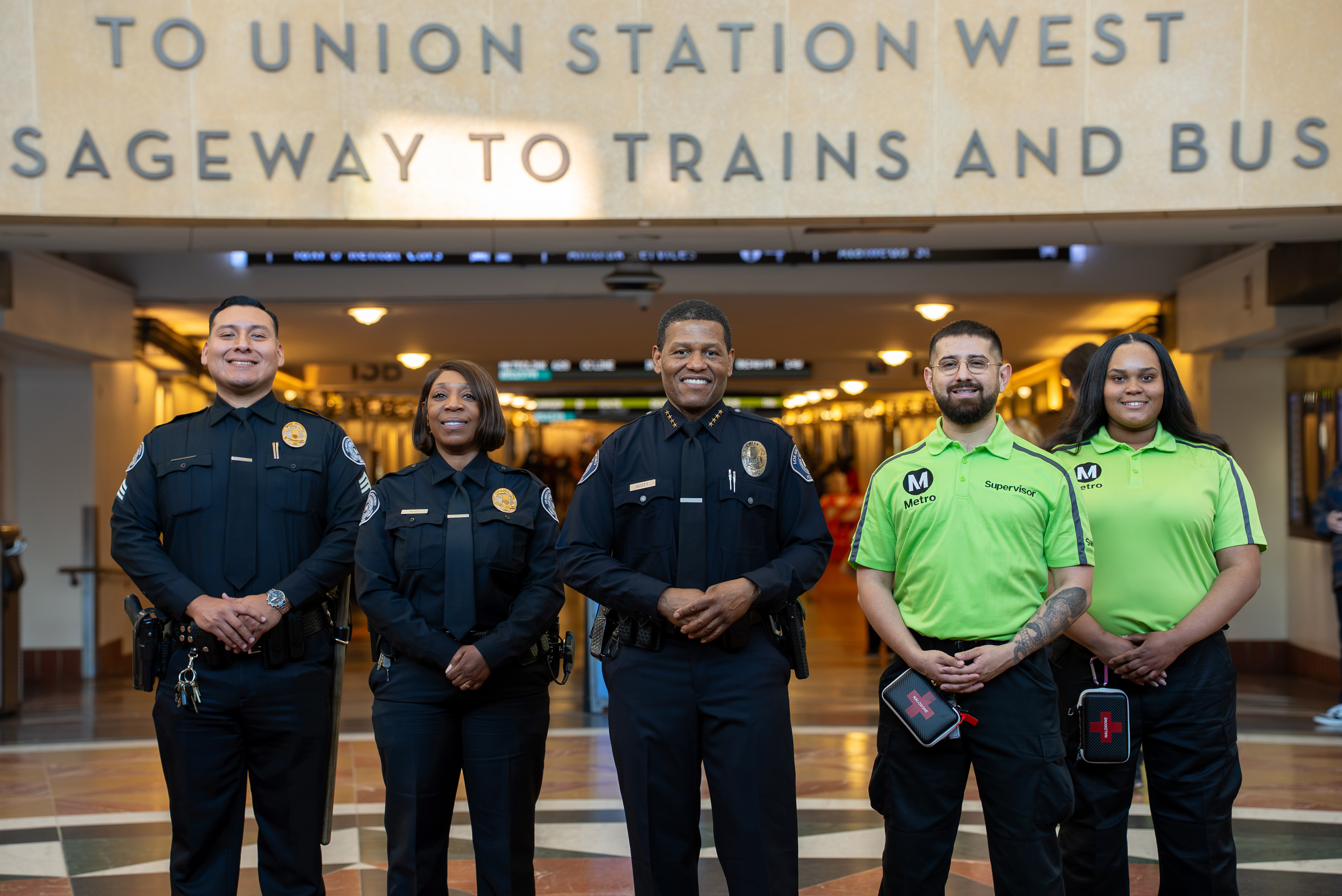 Five uniformed public safety officers standing inside a transit station corridor below signage indicating directions to Union Station West and trains and buses.