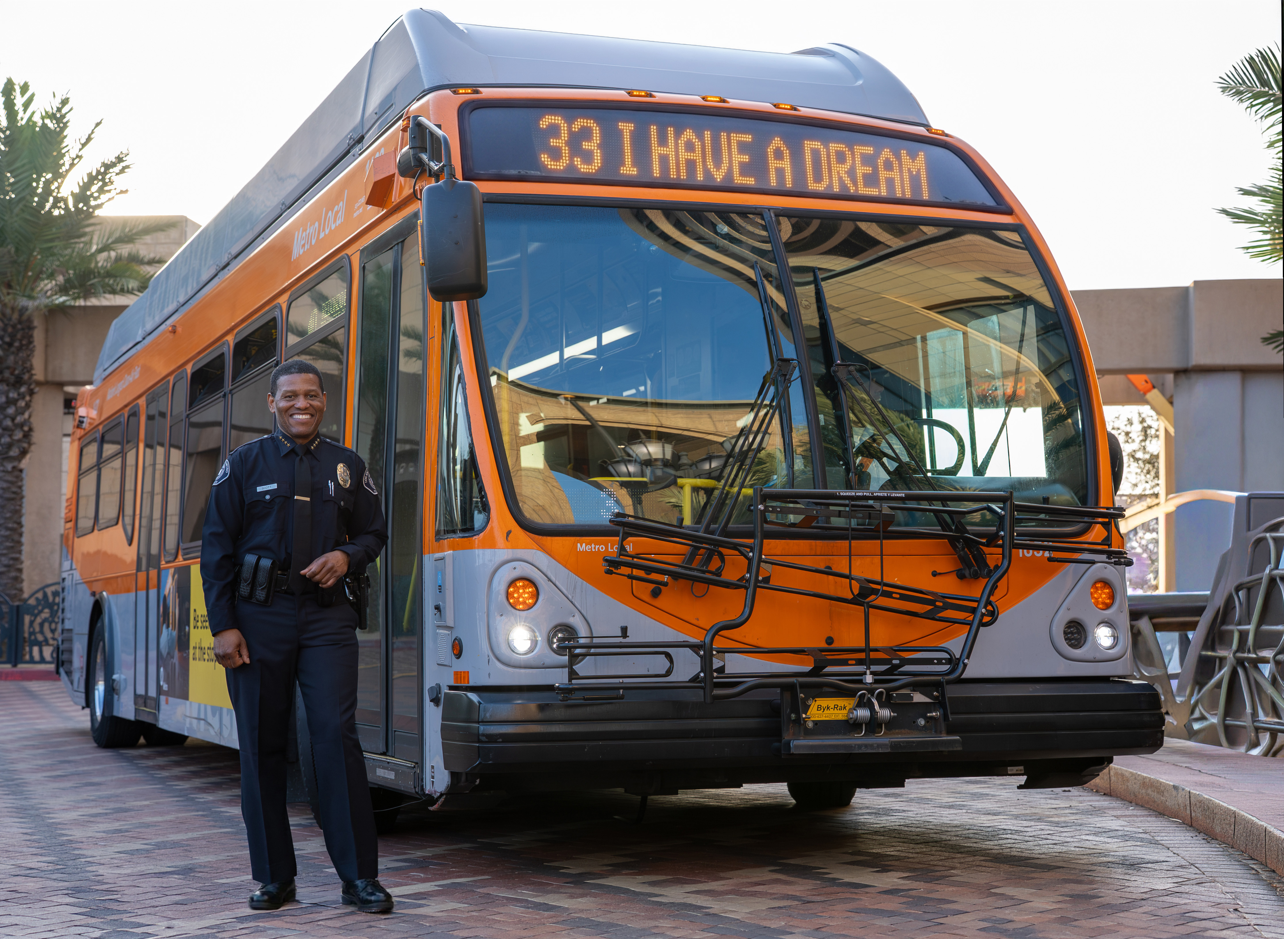 Smiling police officer standing beside an orange Metro Local bus with destination sign reading '33 I HAVE A DREAM'.
