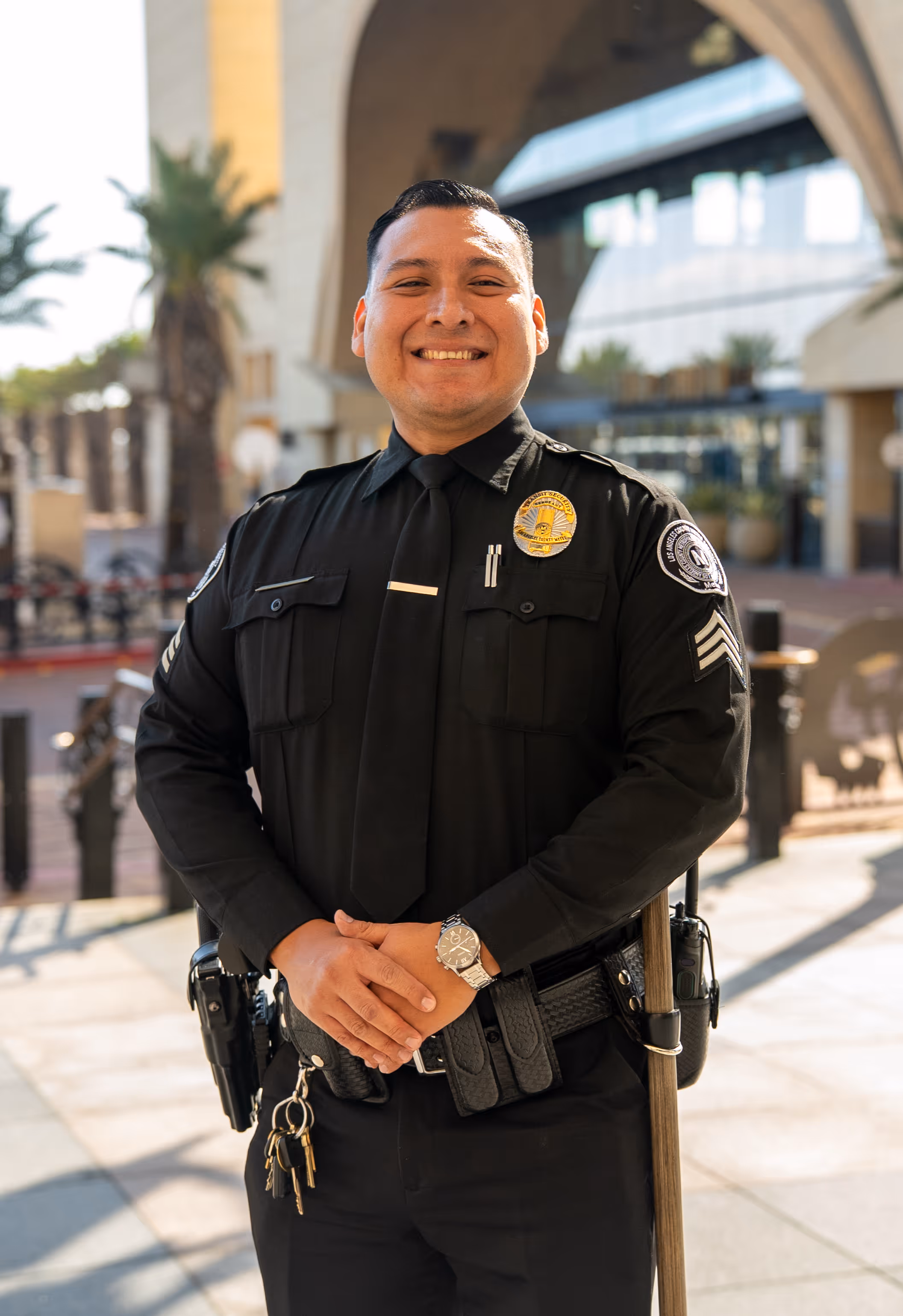 Smiling security officer in black uniform standing outdoors with keys and baton.