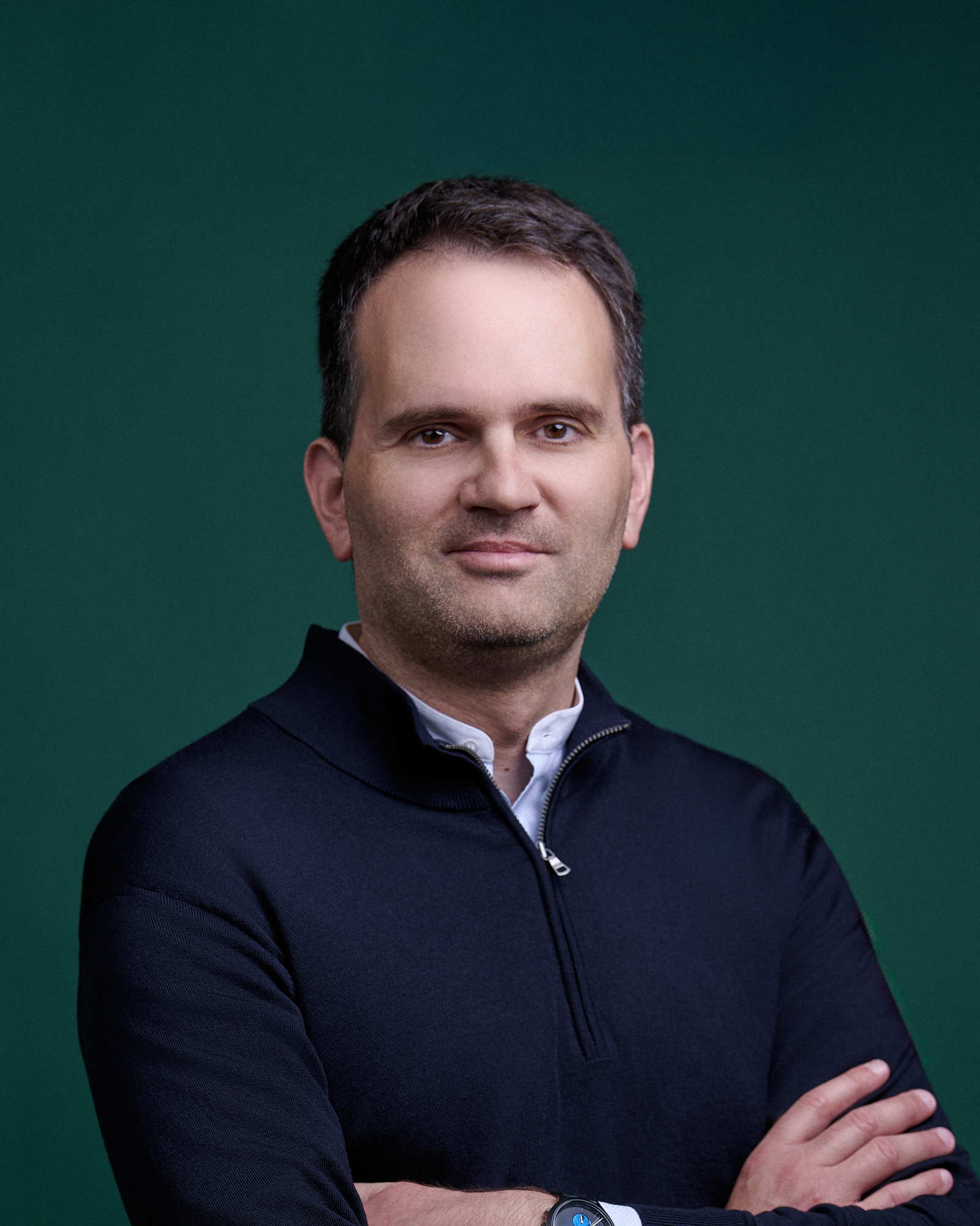 Porträt eines Mannes mit dunkelblauem Pullover und weißem Hemd vor grünem Hintergrund.