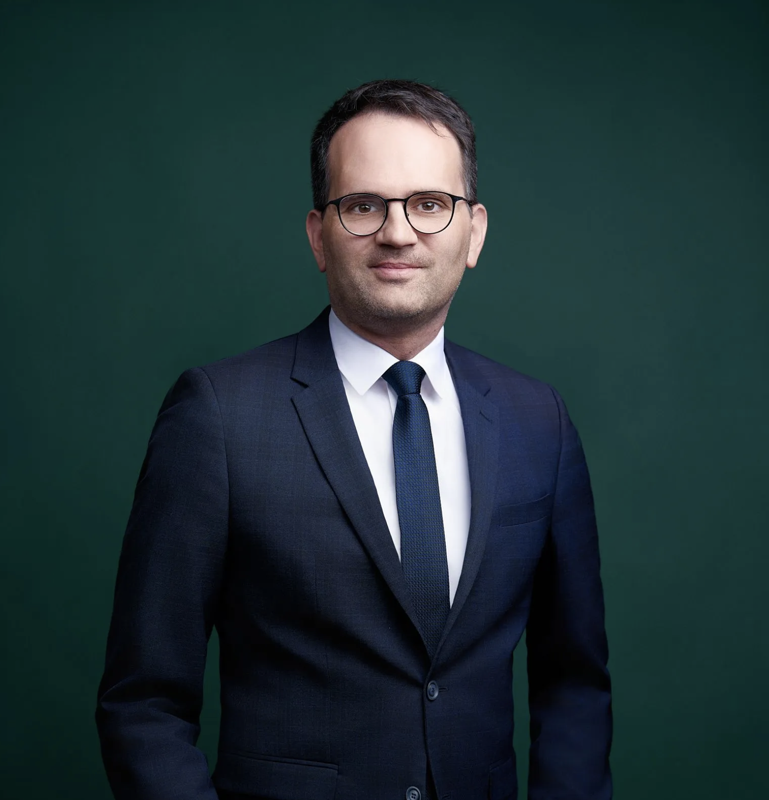Mann trägt dunklen Anzug, weiße Hemd und Krawatte vor grünem Hintergrund.