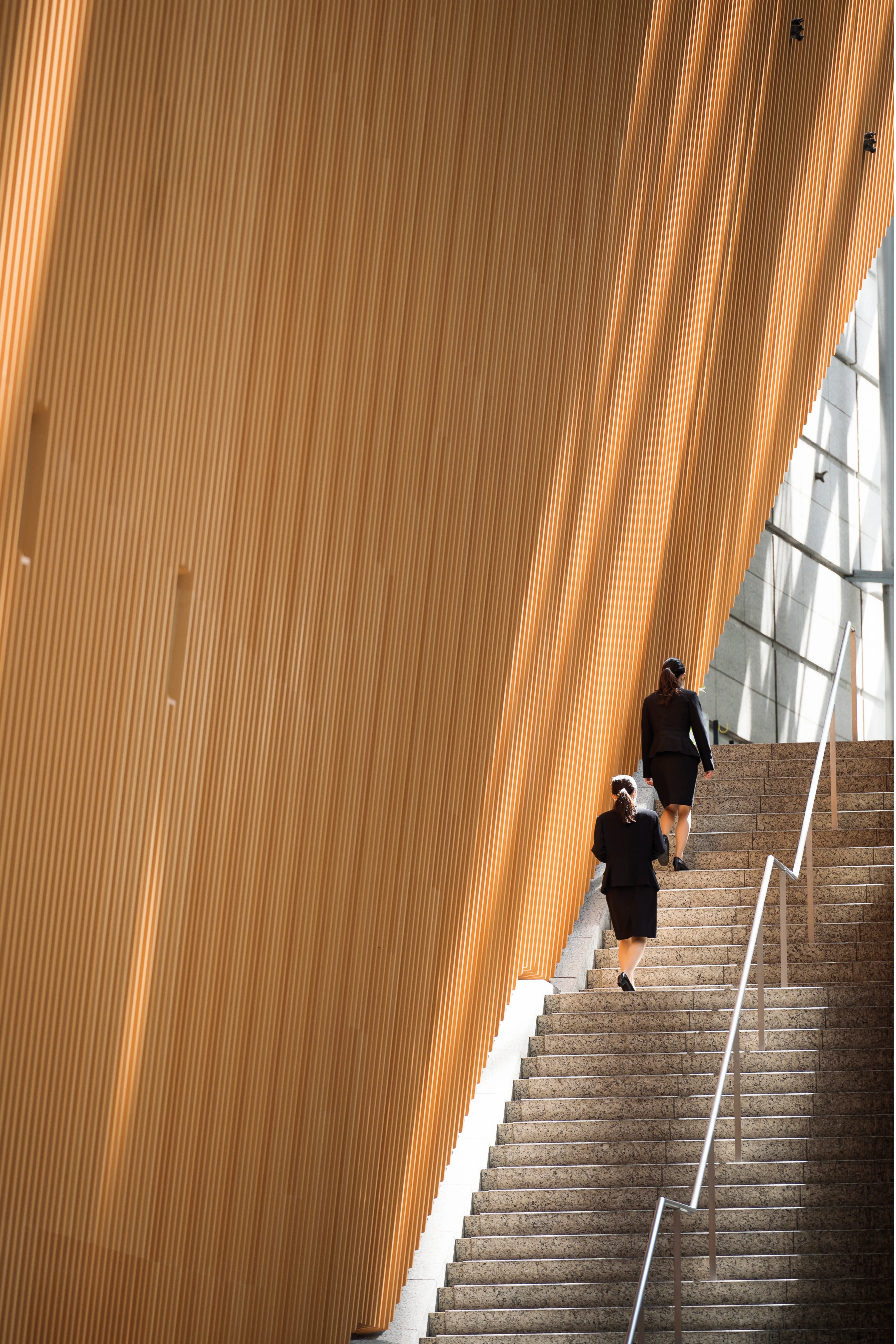 Zwei Frauen in formeller Kleidung steigen eine breite Treppe neben einer großen, vertikal gerillten Holzstruktur hinauf.