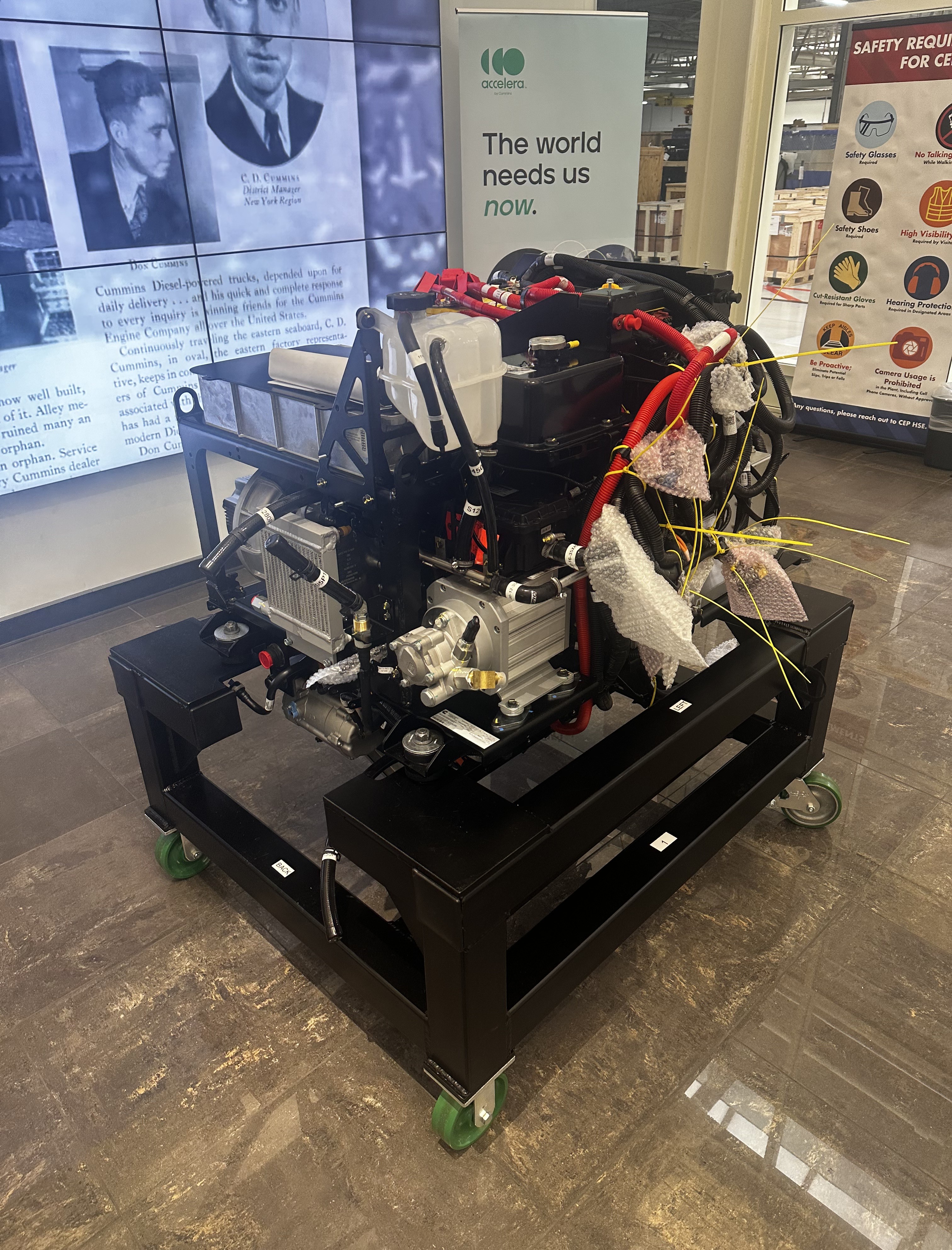 Industrial engine mounted on a black metal frame with green wheels, surrounded by informational banners in a workshop.