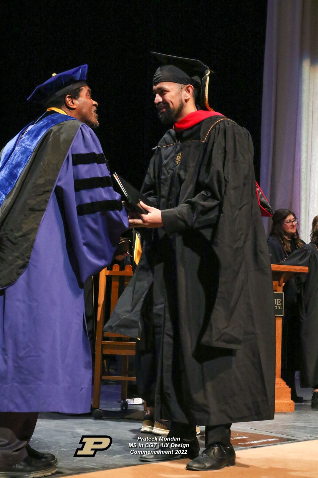 Graduate in black cap and gown with red hood shakes hands with official in blue academic regalia during commencement ceremony.