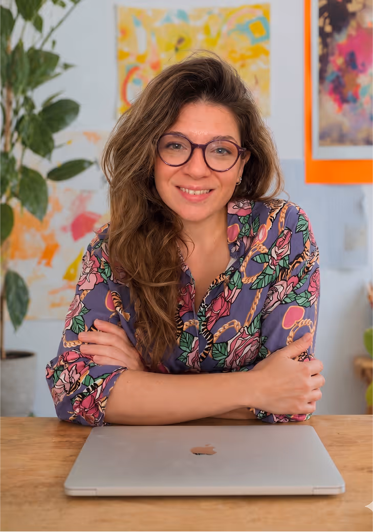 Me with glasses and a messy bun wearing an orange shirt sitting at a desk with art supplies and colorful paintings in the background.