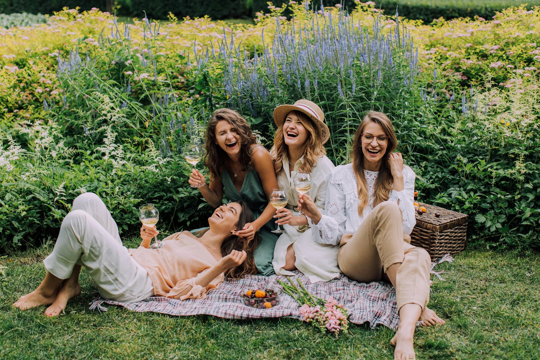 Four women laughing and drinking white wine on a picnic blanket in a garden with flowers and greenery.