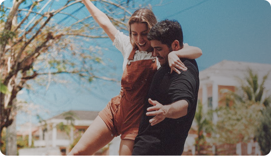 Man in black shirt lifting and playing with a smiling woman in brown overalls outdoors on a sunny day.