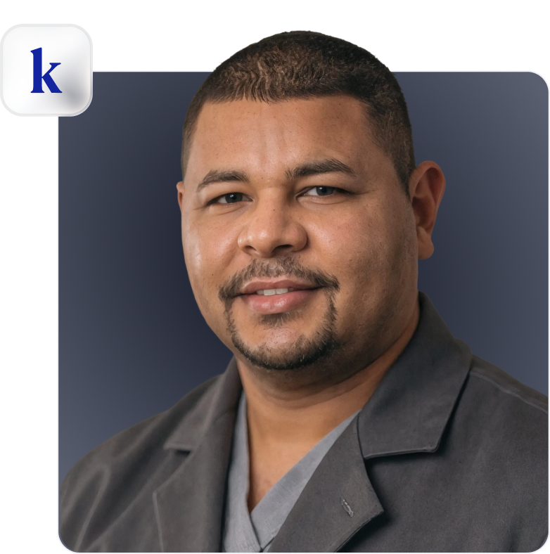 Smiling man with short hair and a trimmed beard wearing a gray shirt and dark jacket on a dark blue background.
