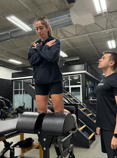 Woman performing back extension exercise on a Roman chair in a gym, with a male trainer observing her form. Ascension Rehab & Sports Therapy