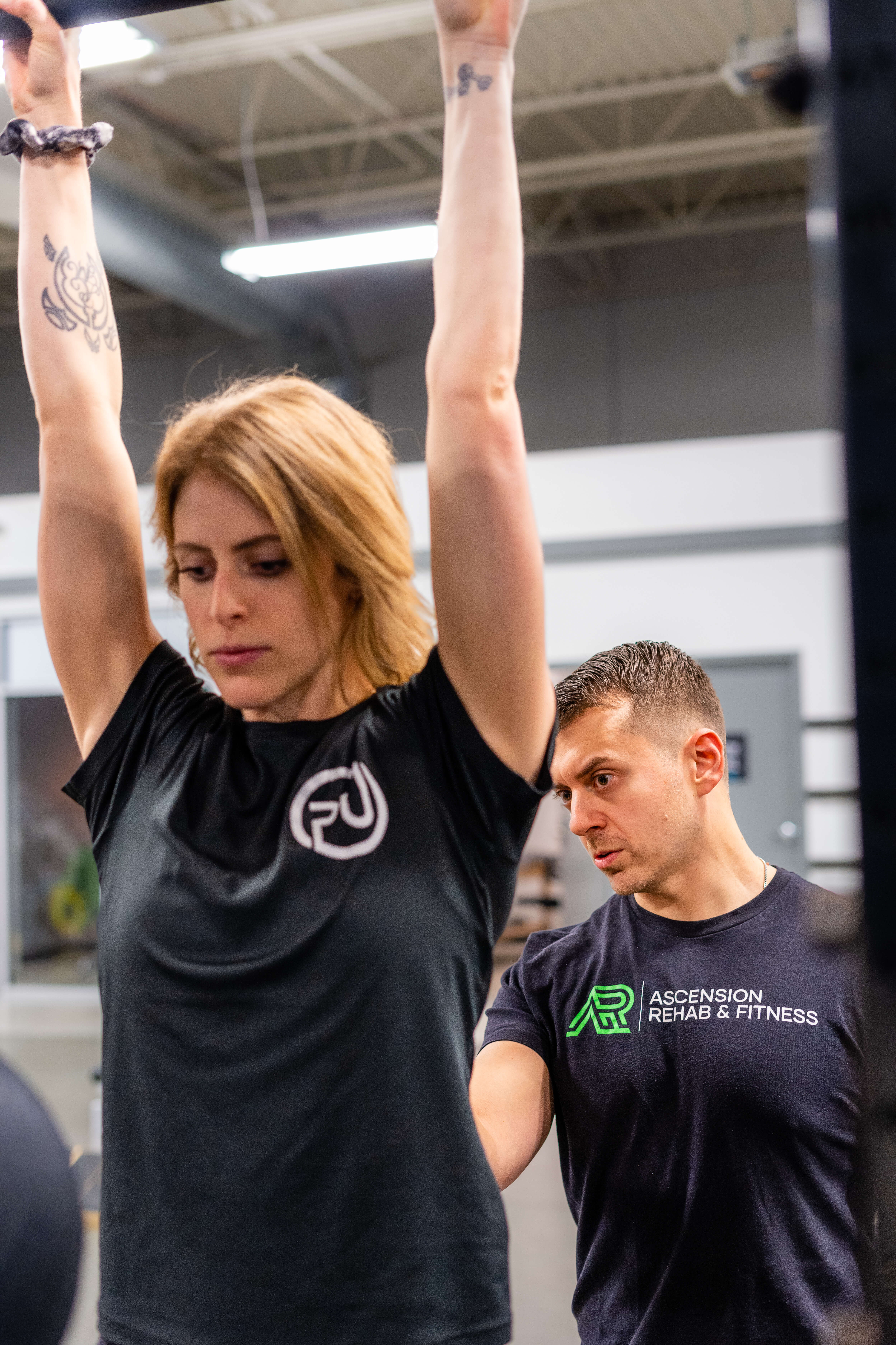 Woman lifting bar overhead during workout while male trainer from Ascension Rehab & Fitness closely observes her form. Ascension Rehab & Sports Therapy