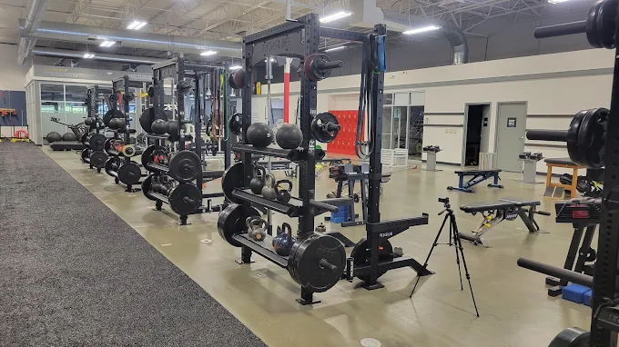 Spacious gym interior with multiple squat racks equipped with weights, kettlebells, benches, and resistance bands. Ascension Rehab & Sports Therapy