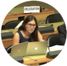 Mujer con gafas y cabello largo sentado en una sala de conferencias con dos computadoras portátiles frente a ella y un cartel que dice delegación.