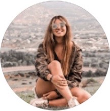 Joven sentada al aire libre con gafas de sol, con un paisaje montañoso desenfocado de fondo.