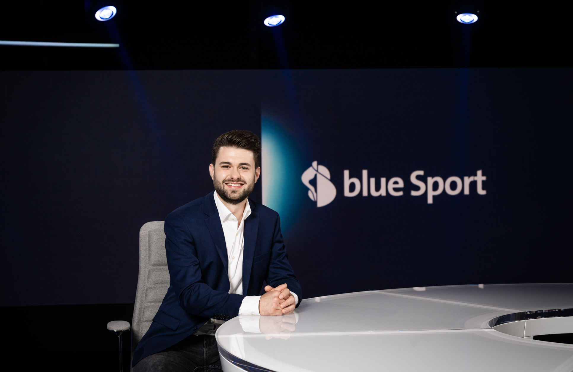 Manuel Iseli im dunkelblauen Anzug sitzt an einem modernen weissen Tisch vor einem Bildschirm mit dem Logo und Schriftzug blue Sport.