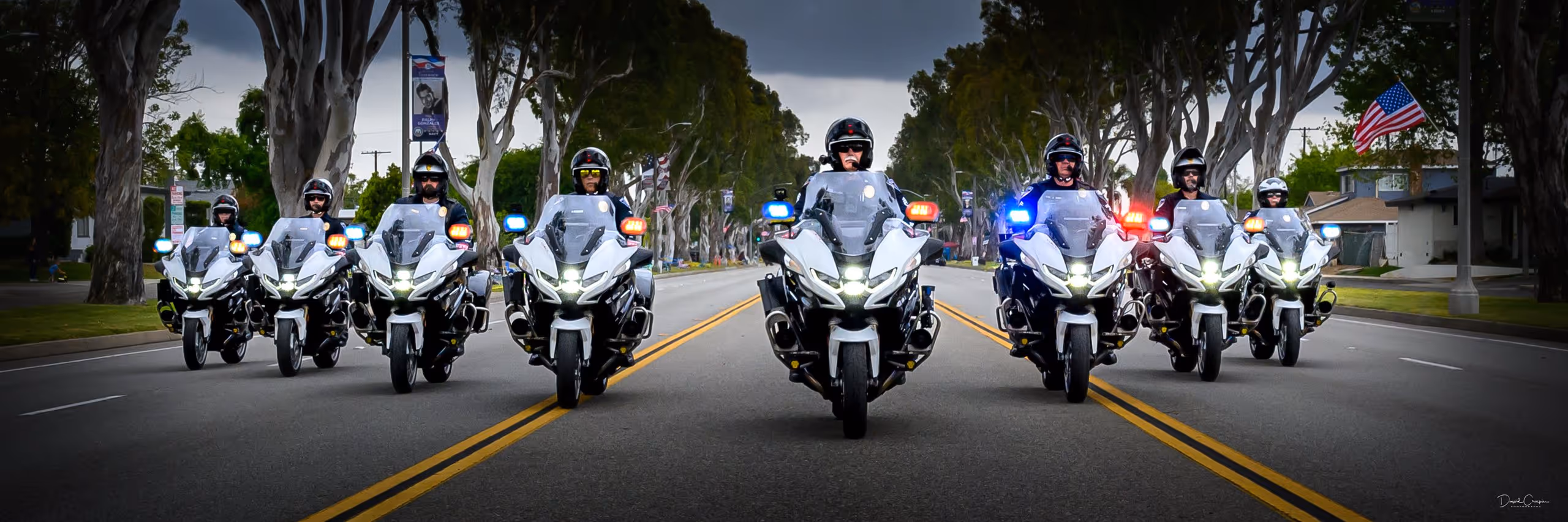 Eight police officers riding white motorcycles with flashing lights, lined up in formation on a tree-lined street.