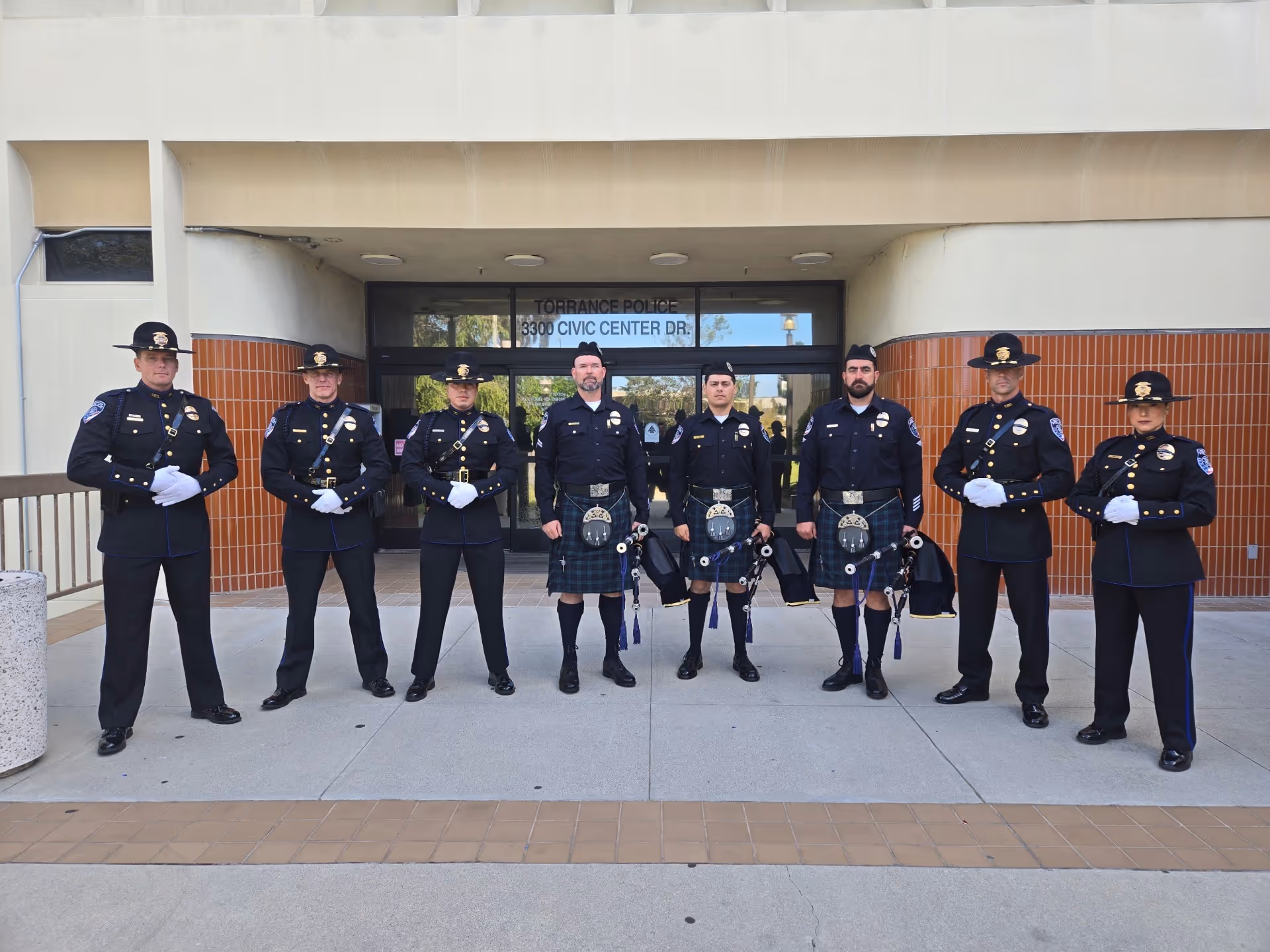 Eight police officers in uniform standing in a row in front of Torrance Police Civic Center entrance, three wearing kilts and holding bagpipes.