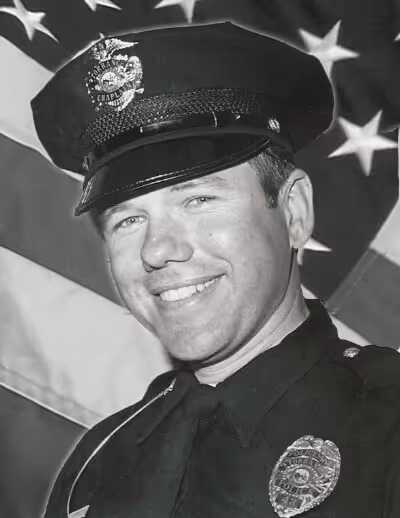 Black and white portrait of a smiling police officer in uniform with an American flag in the background.