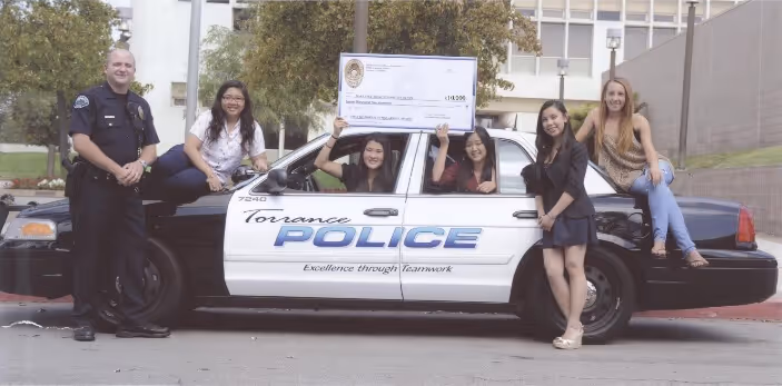 Five young women and a police officer pose with a large check while standing and sitting on a Torrance Police car.