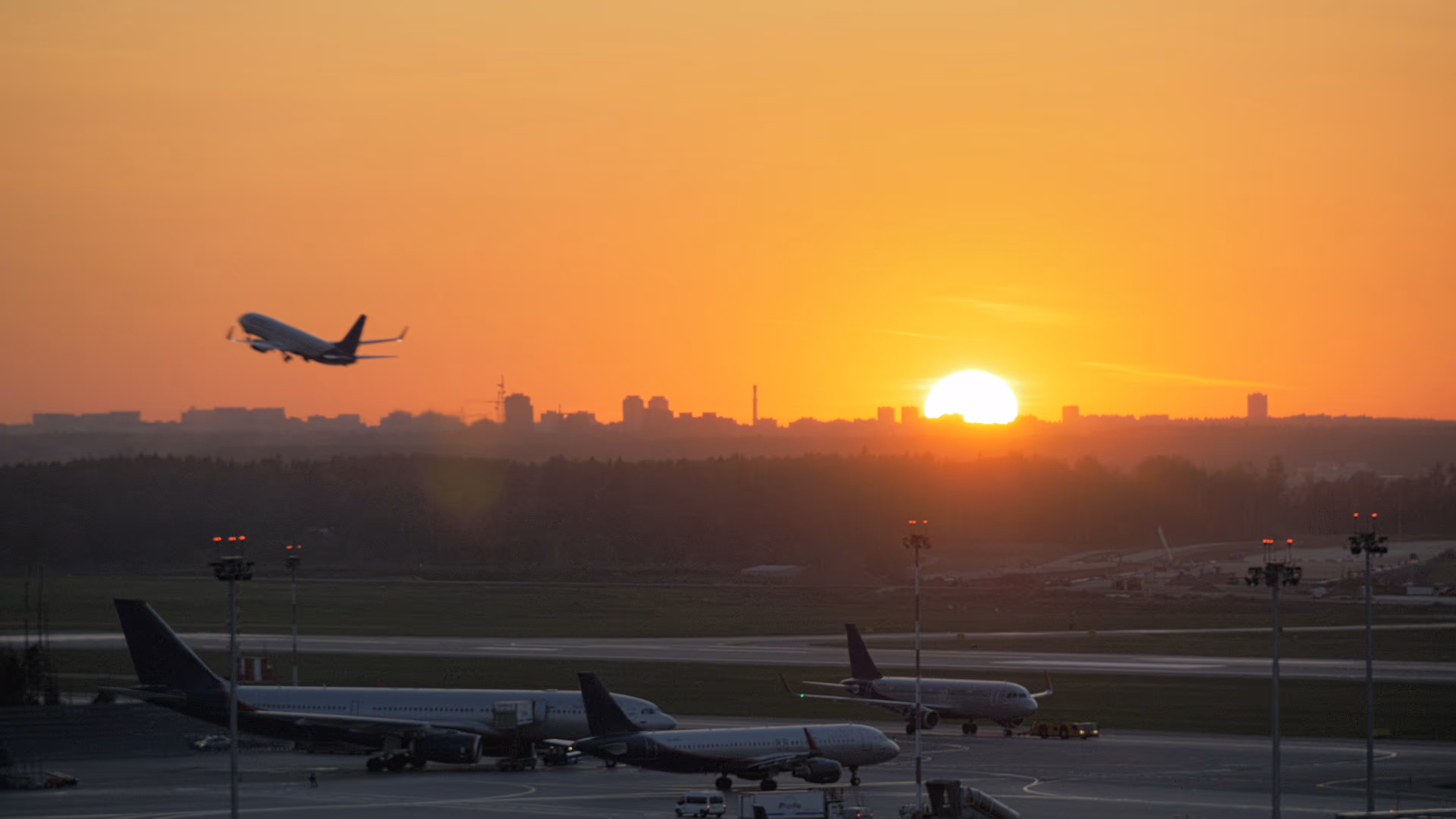Airplane taking off at sunset with other planes parked on runway and city skyline in background.