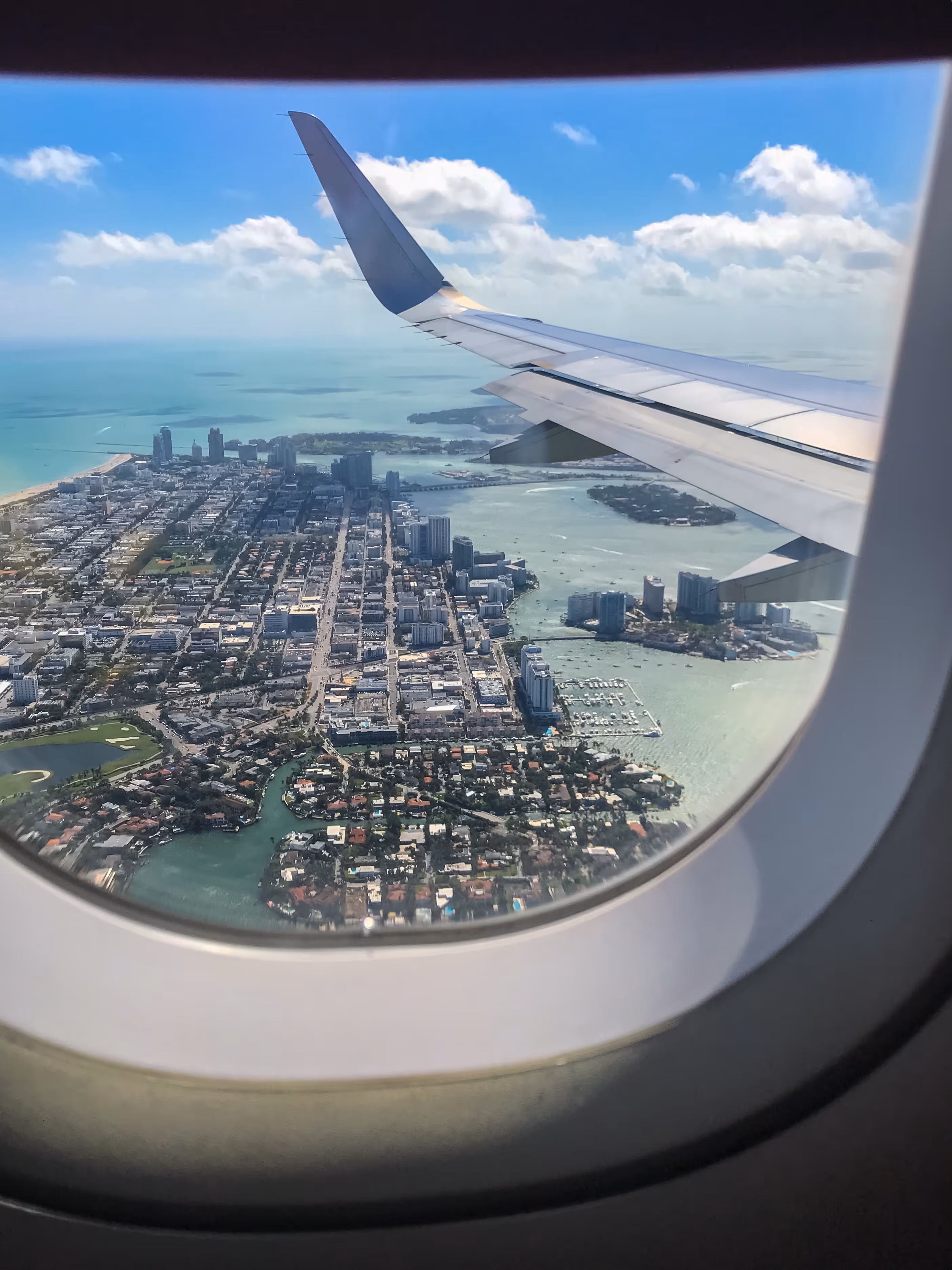 View of a coastal city with buildings and waterways seen through an airplane window with a wing visible.